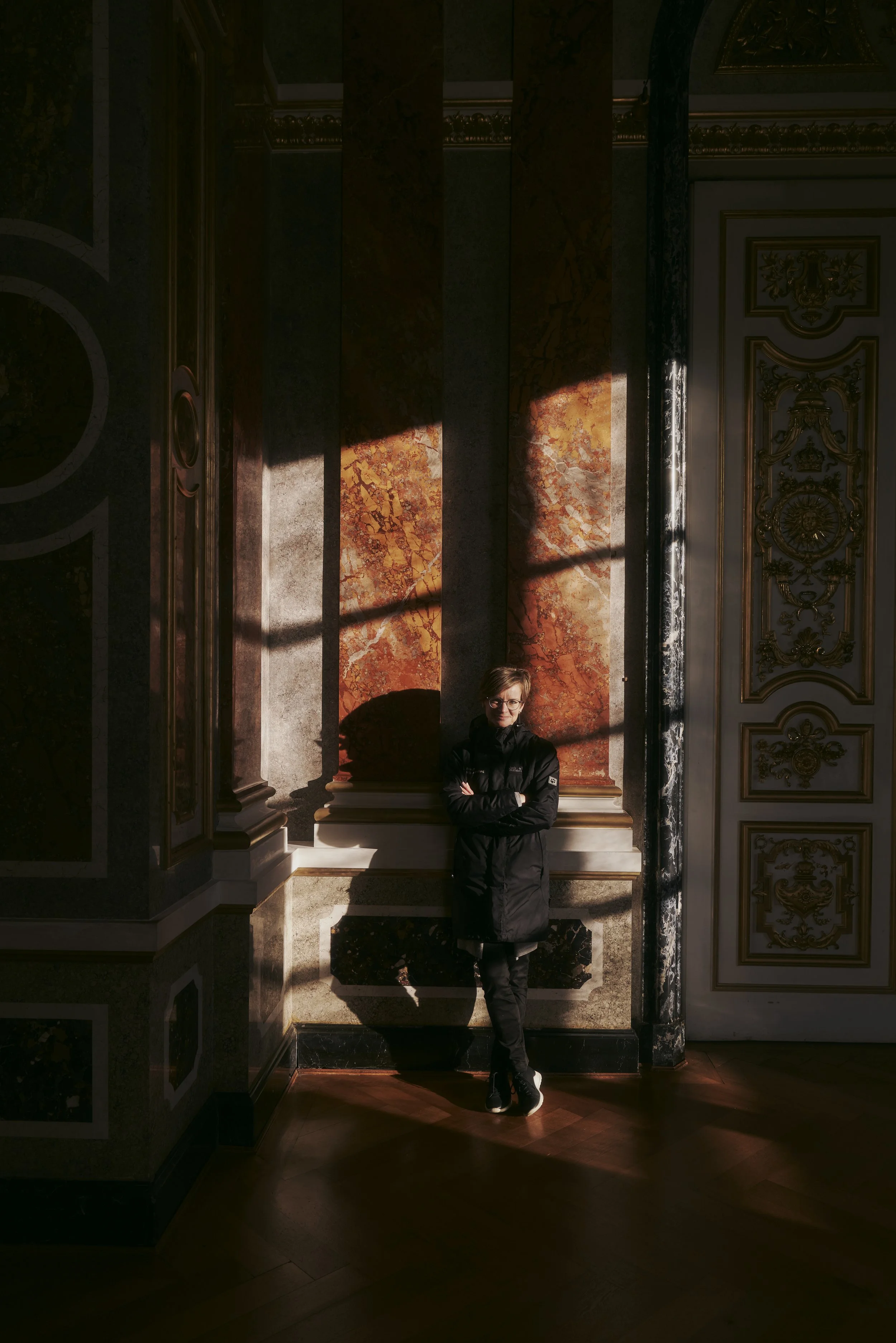 Person steht in einem prunkvoll dekorierten Raum mit goldenen Verzierungen und Marmorsäulen. Sonnenstrahlen fallen auf die Wand hinter der Person.