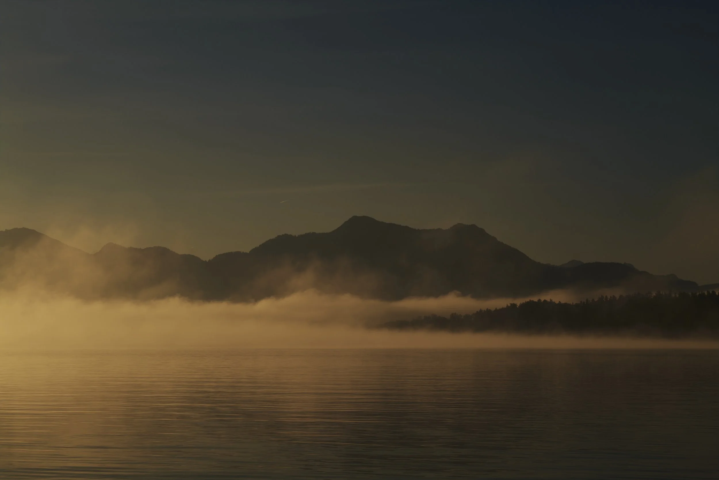 Morgennebel über einem See mit Bergen im Hintergrund, im sanften Licht der Morgendämmerung.