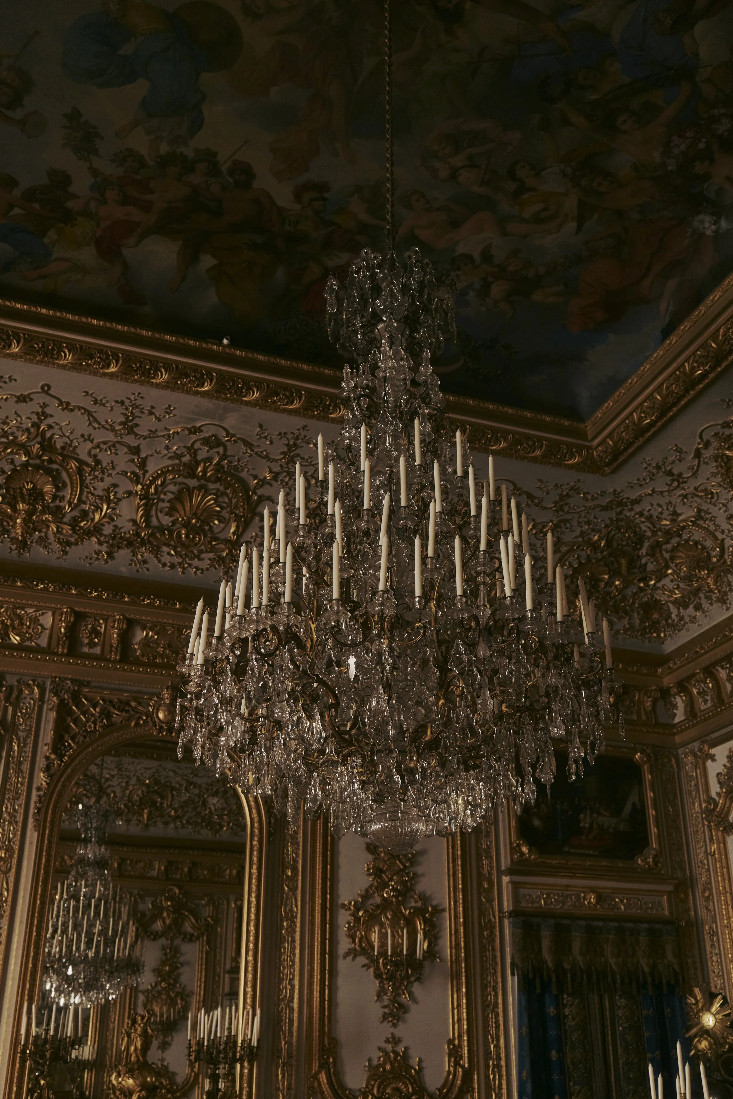 Ein opulenter Kristallkronleuchter in einem reich verzierten Raum mit goldenen Stuckverzierungen und einem bemalten Deckenfresko.