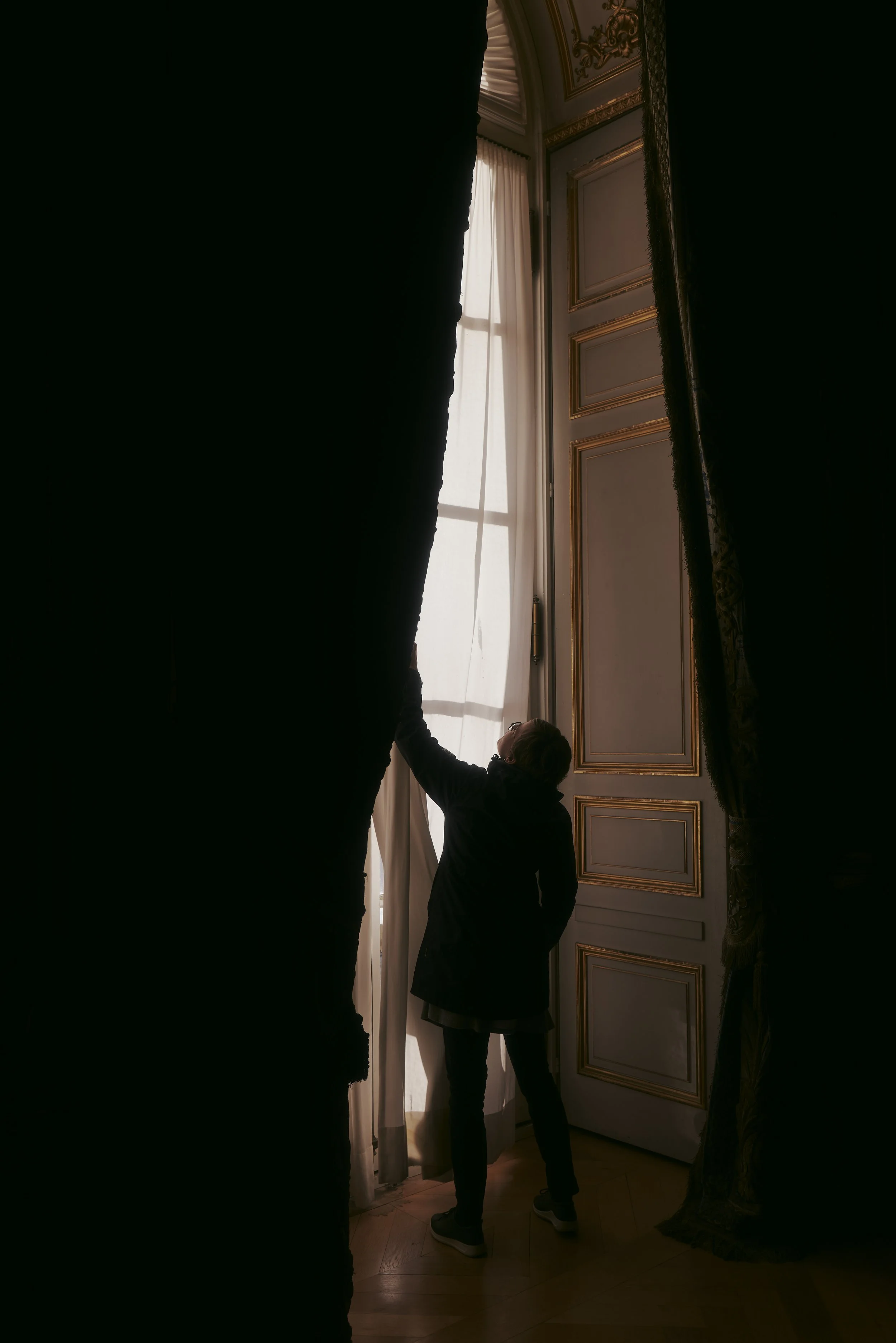 Person zieht einen Vorhang in einem elegant dekorierten Raum auf, mit hohen Decken und großen Fenstern, die mit weißen Gardinen bedeckt sind.