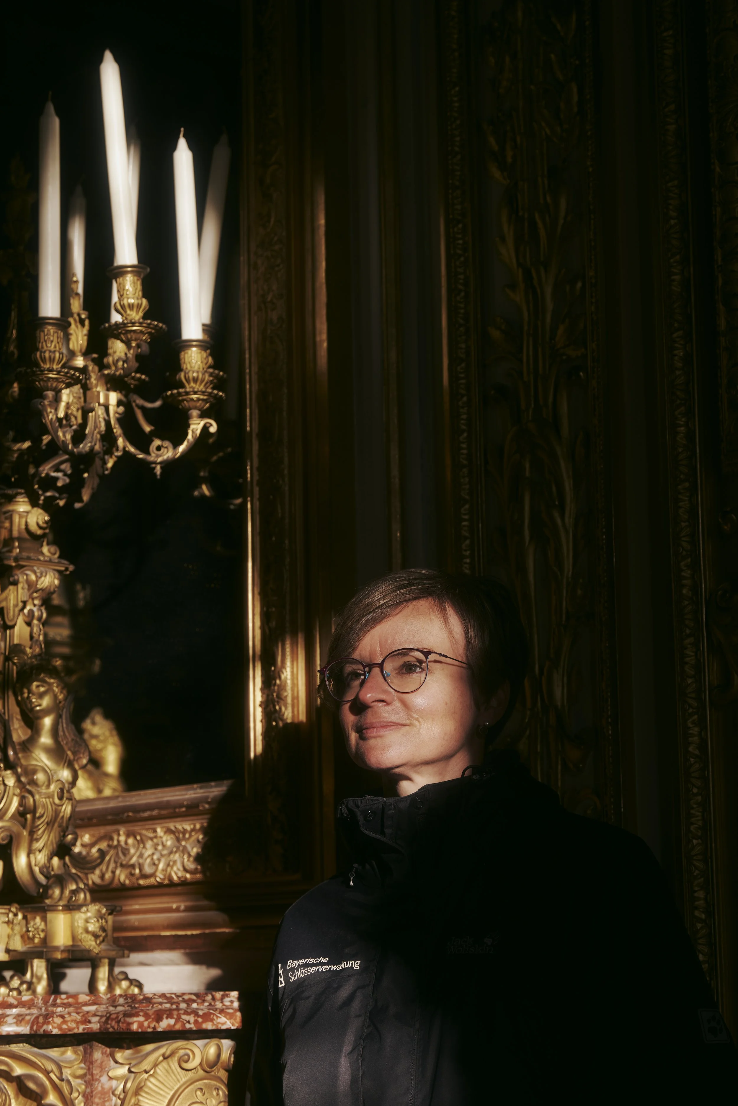 Eine Person steht vor einem goldenen, verzierten Spiegel und einem Kronleuchter mit langen Kerzen. Die Person trägt eine schwarze Jacke mit der Aufschrift "Bayerische Schlösserverwaltung". Es ist ein kunstvoll gestalteter Raum mit dekorativen Element