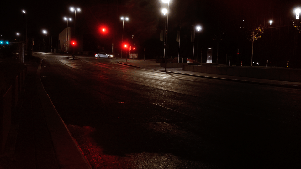 Nächtliche Stadtszene mit leerer Straße, beleuchtet von Straßenlaternen und roten Ampeln. Ein vorbeifahrendes Auto ist sichtbar.