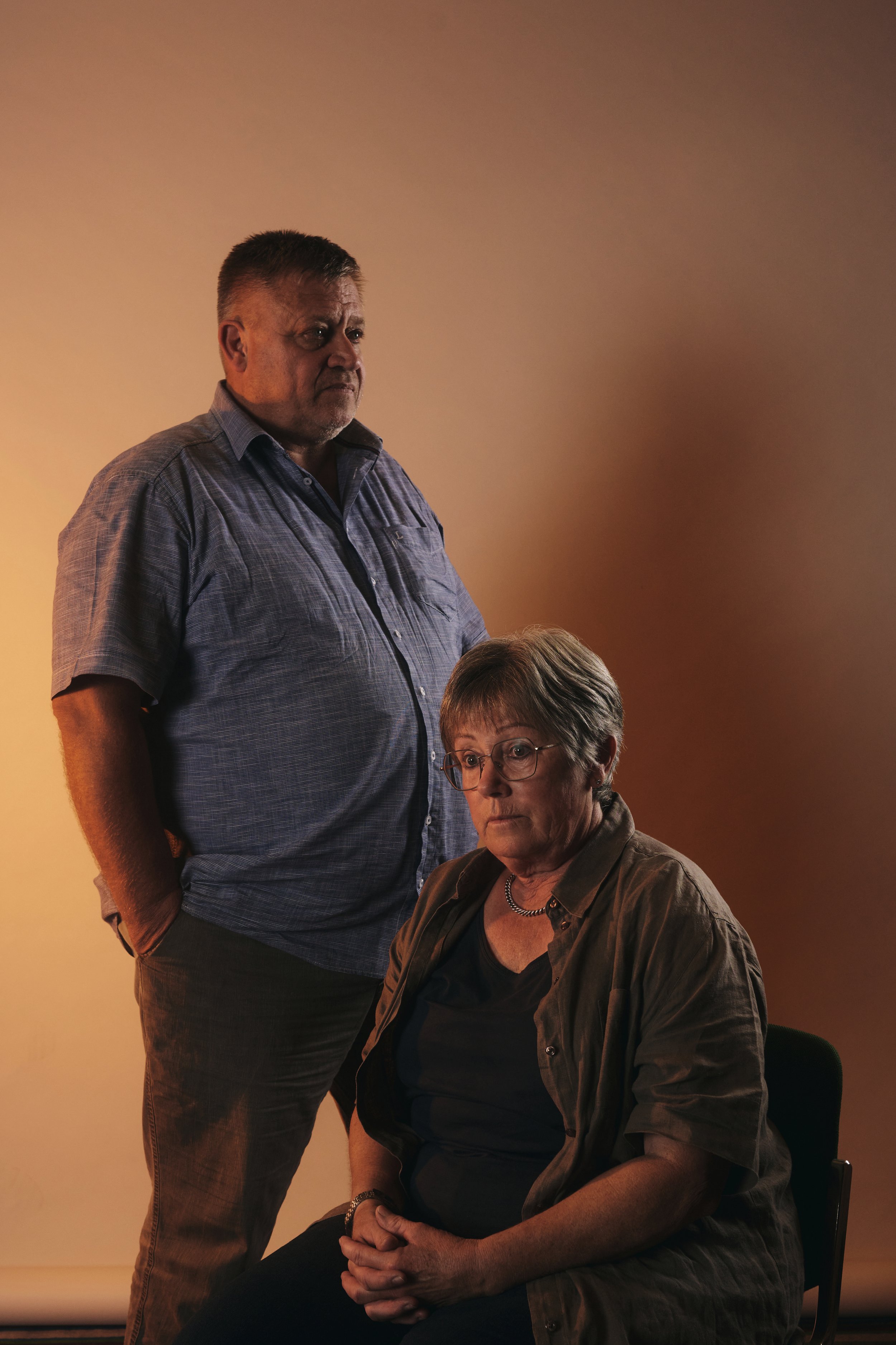 Mann steht und Frau sitzt, vor neutralem Hintergrund.