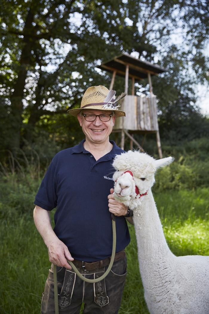 Ein Mann mit Strohhut steht lächelnd neben einem weißen Alpaka in einem grünen Außenbereich mit einem Hochsitz im Hintergrund.