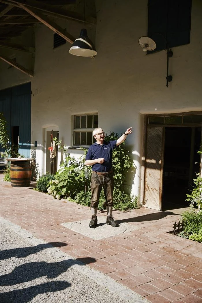 Ein Mann steht vor einem Gebäude mit traditioneller Kleidung, möglicherweise in Lederhosen. Er zeigt auf etwas außerhalb des Bildes. Im Hintergrund sieht man Pflanzen, einen Holztürrahmen und ein Holzfass. Die Szene ist sonnig und wirkt ländlich.