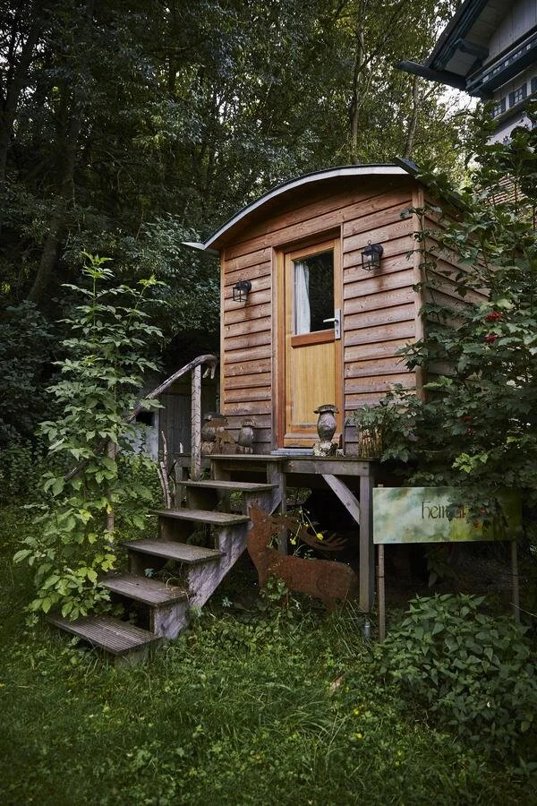 Kleines Holzhaus im Garten mit Treppe, umgeben von Bäumen und Pflanzen im Grünen.