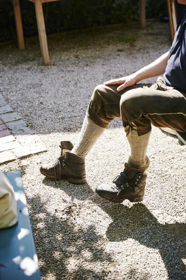 Person sitzt im Freien und trägt traditionelle Lederhose, dicke Wollsocken und Wanderschuhe. Ein Schuh ist teilweise geöffnet und die Schnürsenkel sind locker. Kieselboden und Holztisch im Hintergrund.