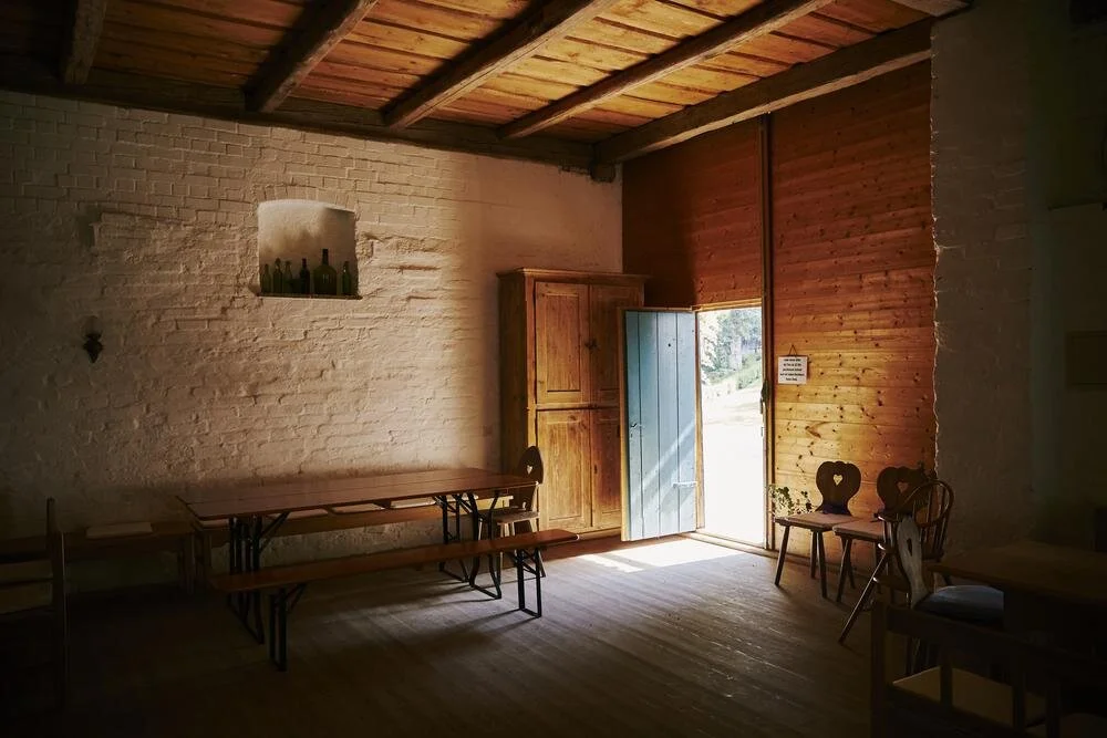 Rustikaler Raum mit Holztisch und Bänken, Holzvertäfelung, offene Tür mit Licht, ein Schrank, und Dekorflaschen auf der Fensterbank. Gemütliche, einfache Einrichtung.