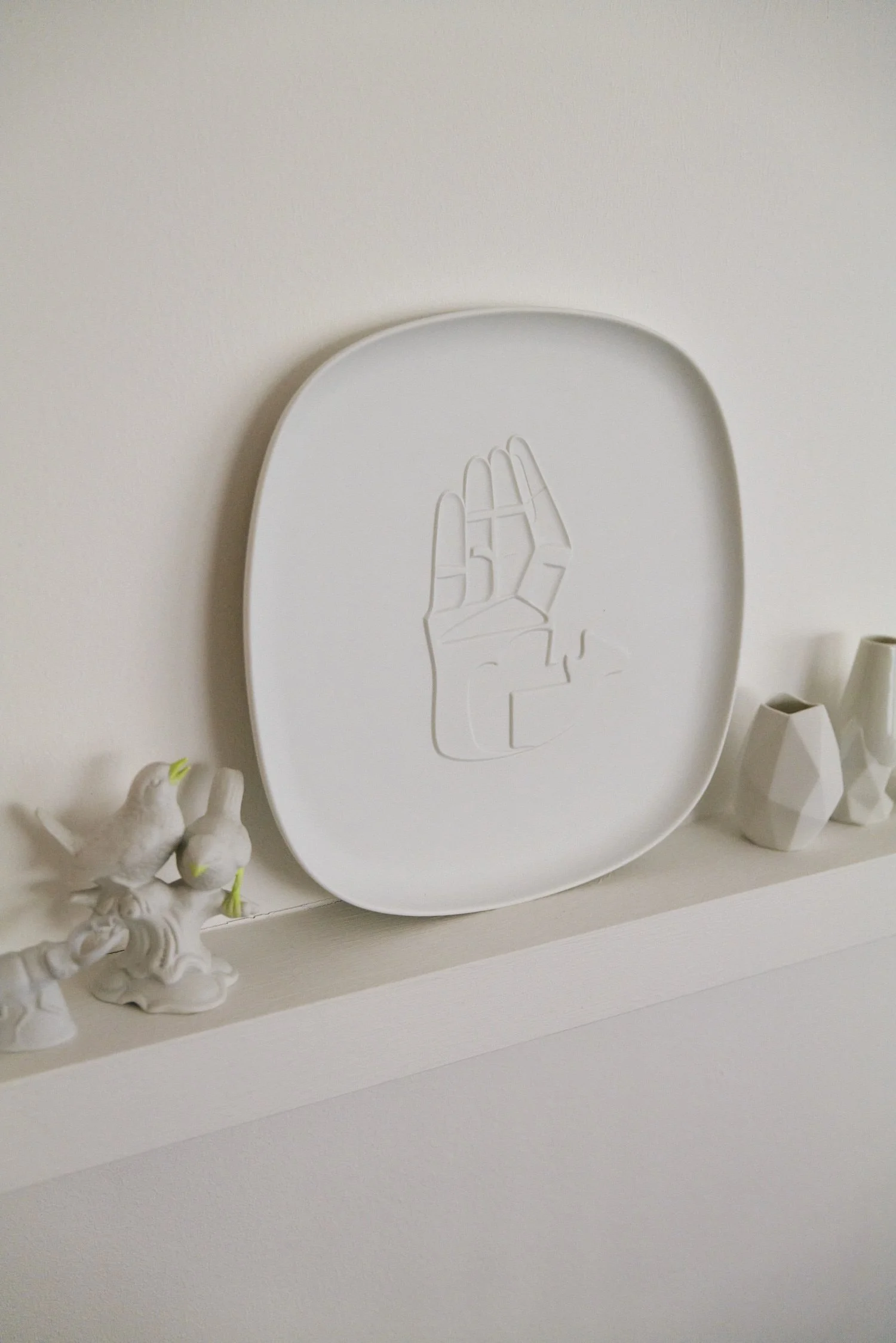 Weißer Teller mit eingeprägtem Handmotiv, daneben weiße Porzellanfiguren von Vögeln und modernen Vasen, auf einem weißen Regal.