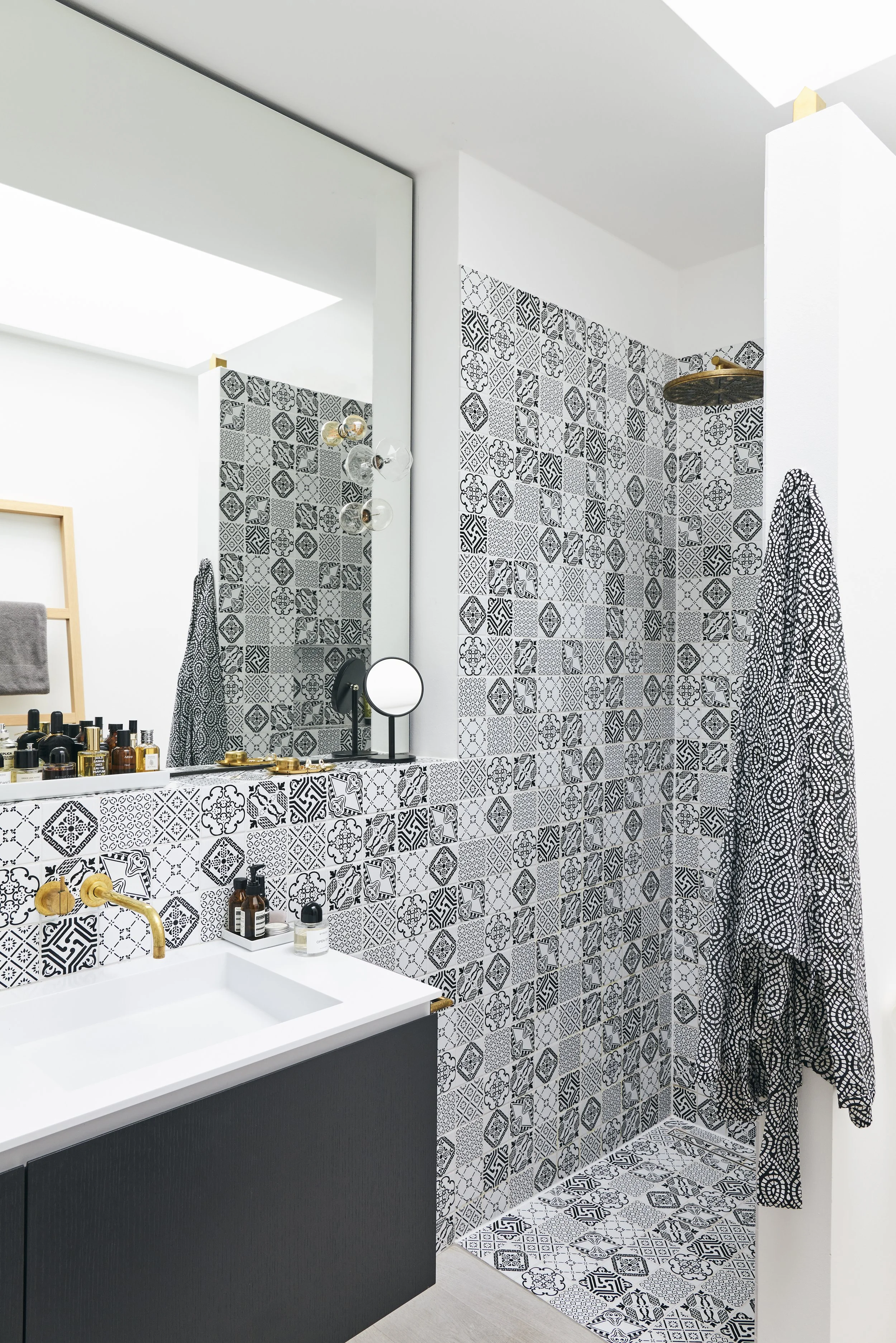 Modernes Badezimmer mit schwarz-weiß gemusterten Fliesen, einem weißen Waschbecken, schwarzen Schränken, einem großen Spiegel und goldenen Armaturen. Duschbereich und Pflegeprodukte vorhanden.