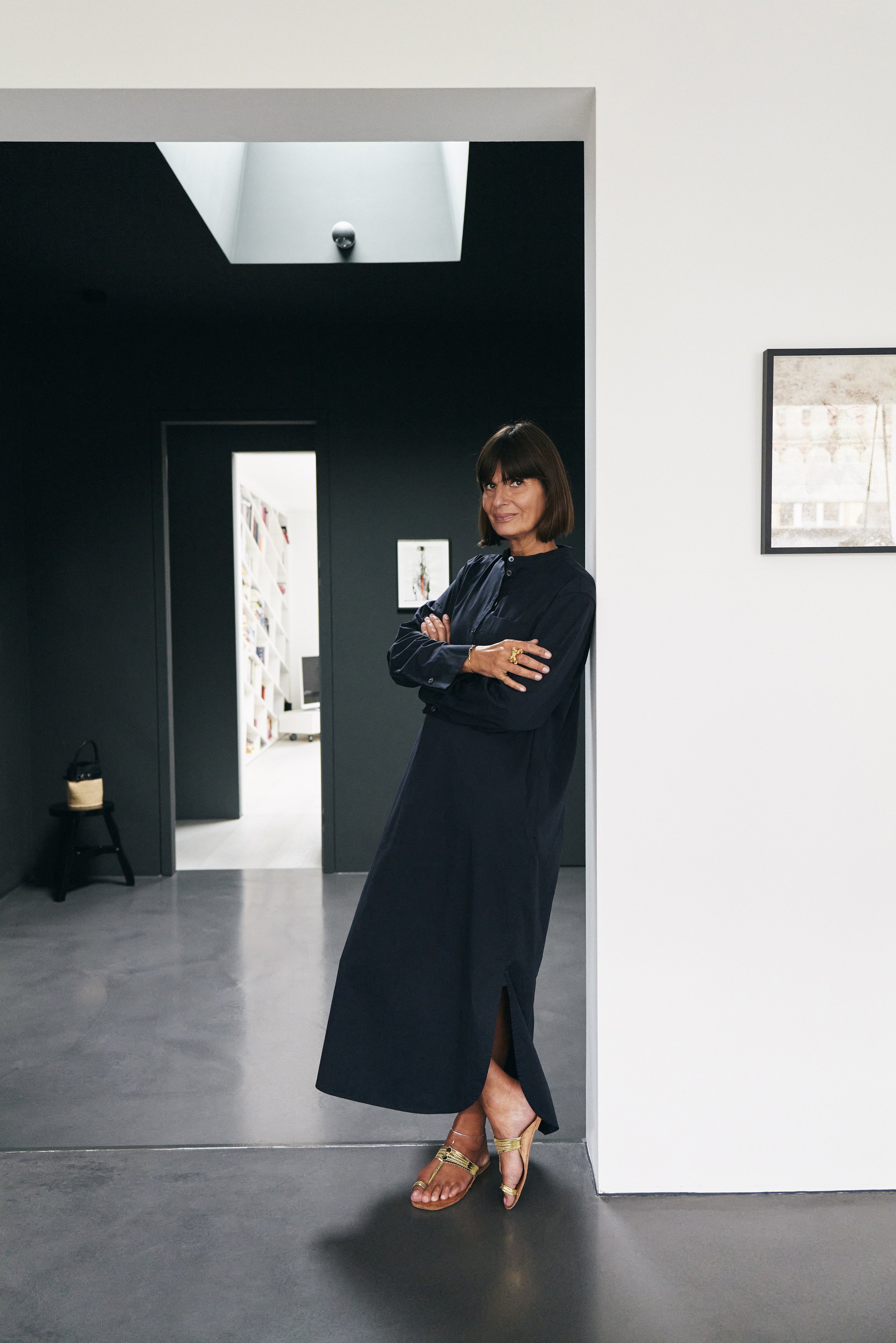 Eine Frau lehnt lächelnd an einer Wand in einem modern eingerichteten Raum, in einem langen schwarzen Kleid, die Arme verschränkt, mit goldenen Sandalen. Im Hintergrund ist ein Durchgang mit hellen Bücherregalen zu sehen.