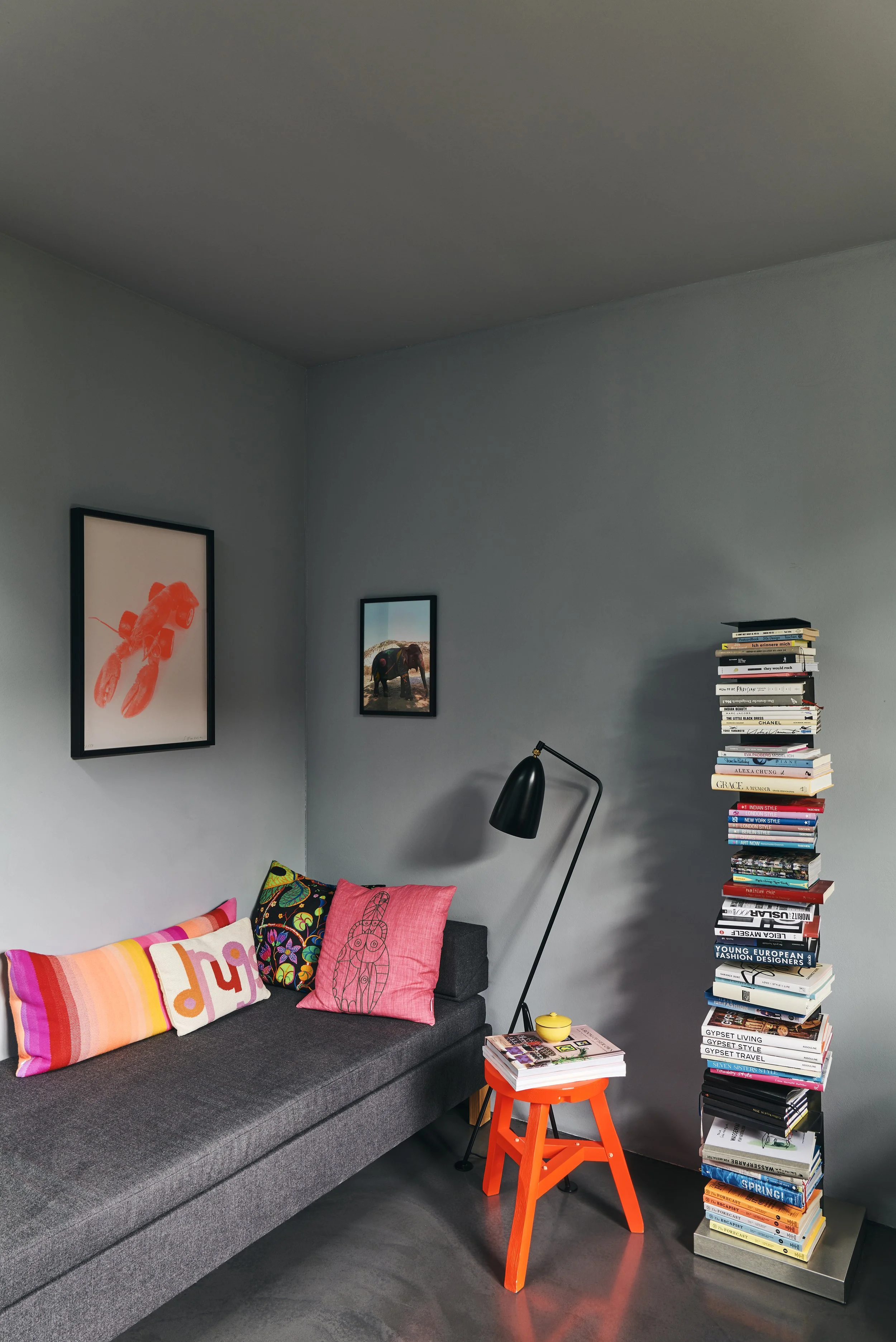 Moderne Sitzecke mit grauem Sofa und bunten Kissen, daneben ein hoher Stapel Bücher. Zwei Wandbilder und eine schwarze Stehlampe sorgen für Dekoration. Ein kleiner, roter Beistelltisch mit weiteren Büchern und einer gelben Tasse steht daneben.
