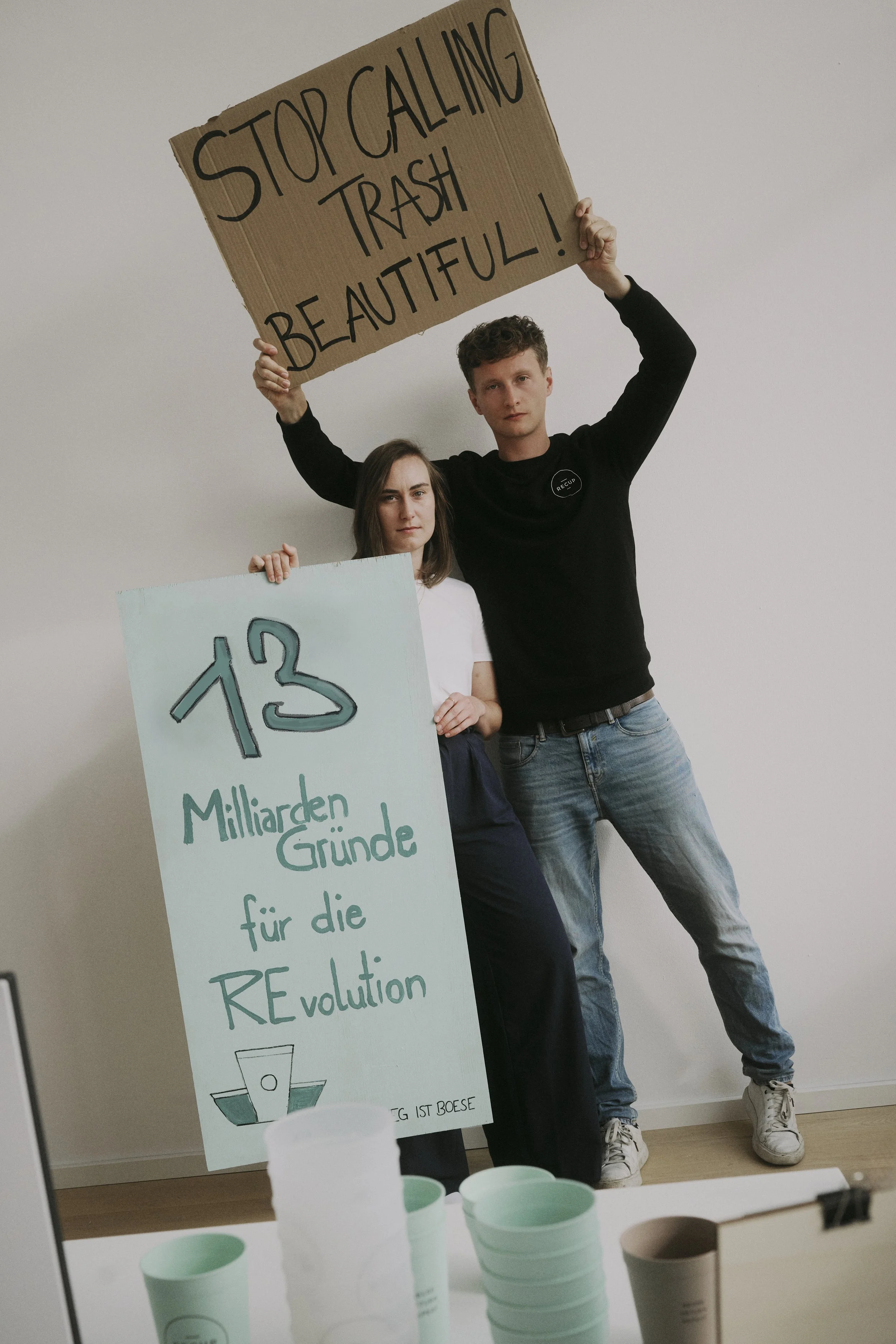 Zwei Personen halten Schilder. Person links hält ein blaues Schild mit der Aufschrift „13 Milliarden Gründe für die Revolution“. Person rechts hält ein braunes Schild mit der Aufschrift „Stop Calling Trash Beautiful!“. Im Vordergrund sind grüne Beche