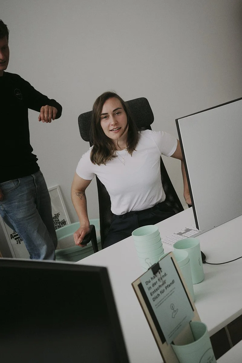 Zwei Personen im Büro, eine sitzt am Schreibtisch vor einem Computerbildschirm, umgeben von Mintgrün-bechern und einem Klemmbrett.