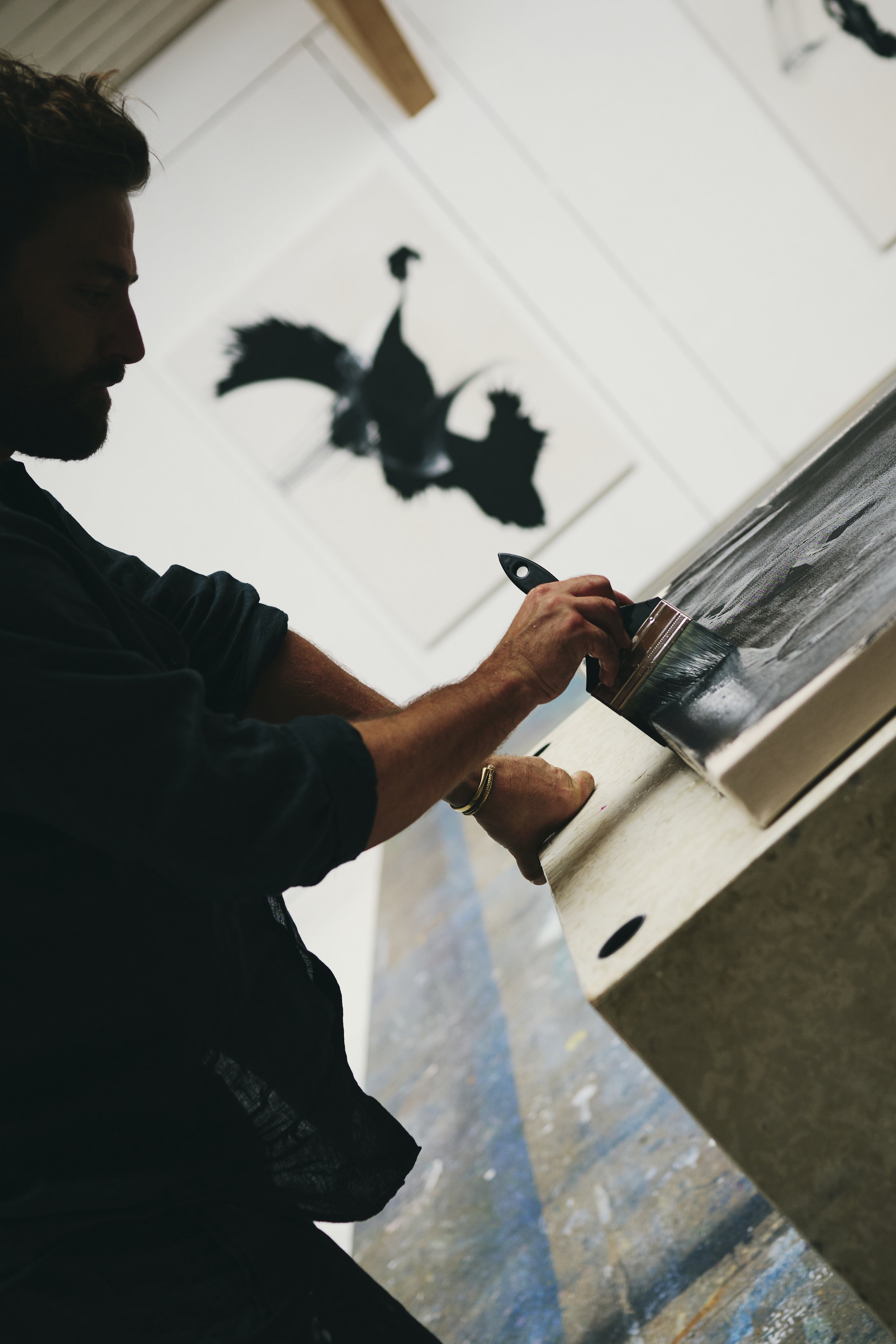 Eine Person malt mit einem Pinsel auf eine Leinwand in einem Atelier; im Hintergrund sind Kunstwerke an der Wand zu sehen.