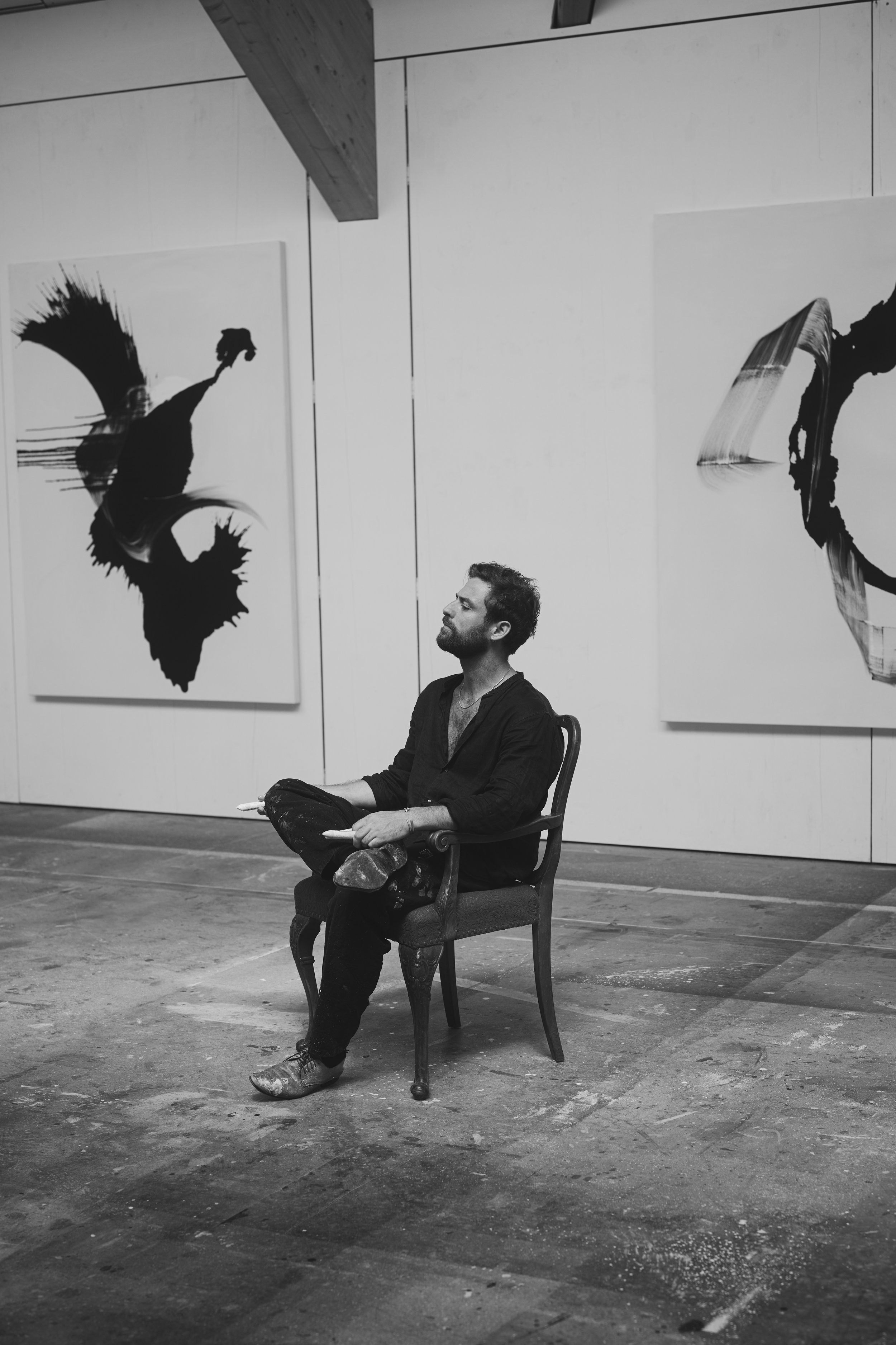 Ein Mann sitzt in einem Kunststudio auf einem Stuhl. Um ihn herum hängen abstrakte Gemälde an der Wand. Die Szene ist in Schwarzweiß gehalten.