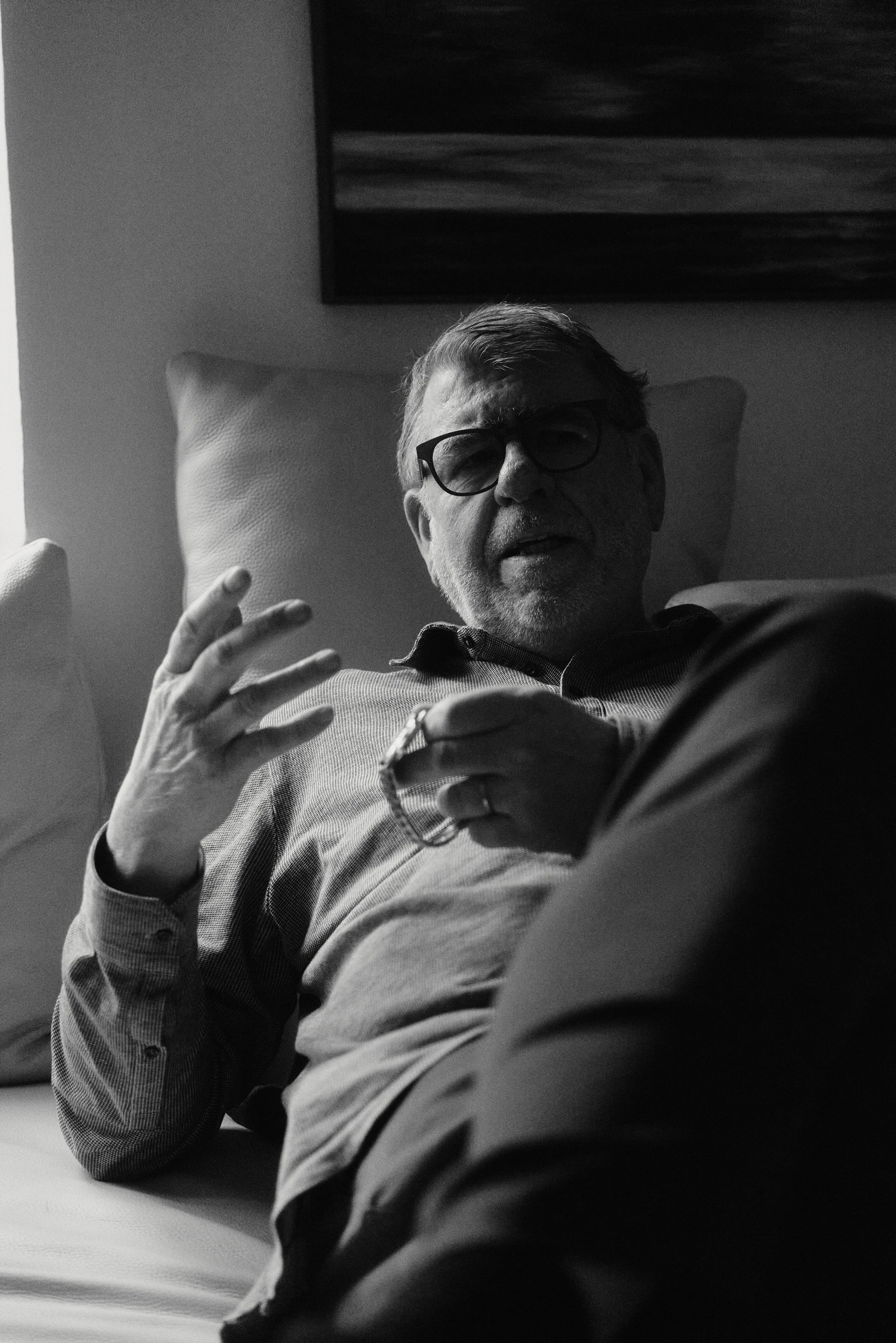 Ein Mann mit Brille und Bart sitzt auf einem Sofa, hält eine Brille und gestikuliert mit der anderen Hand. Schwarz-weiß-Fotografie mit künstlerischer Beleuchtung.