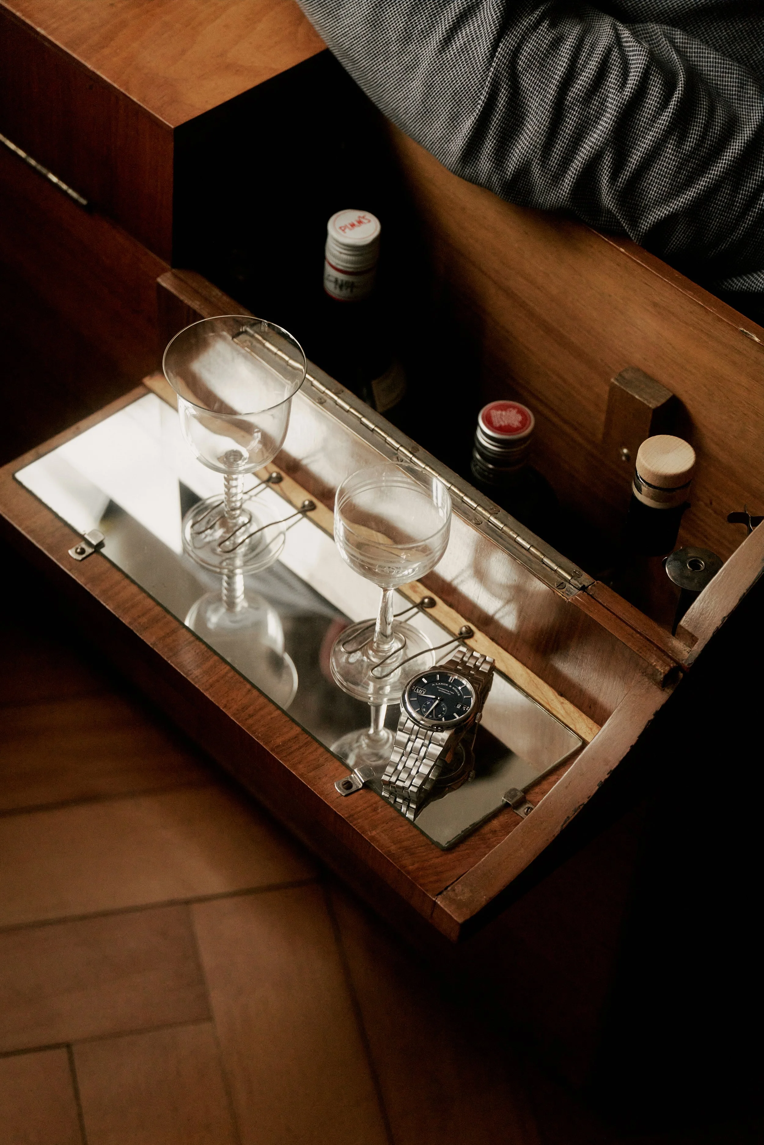 Nahaufnahme eines offenen Holzschranks mit Glasboden, zwei leeren Weingläsern und einer silbernen Uhr, umgeben von mehreren Flaschen mit Verschlüssen.