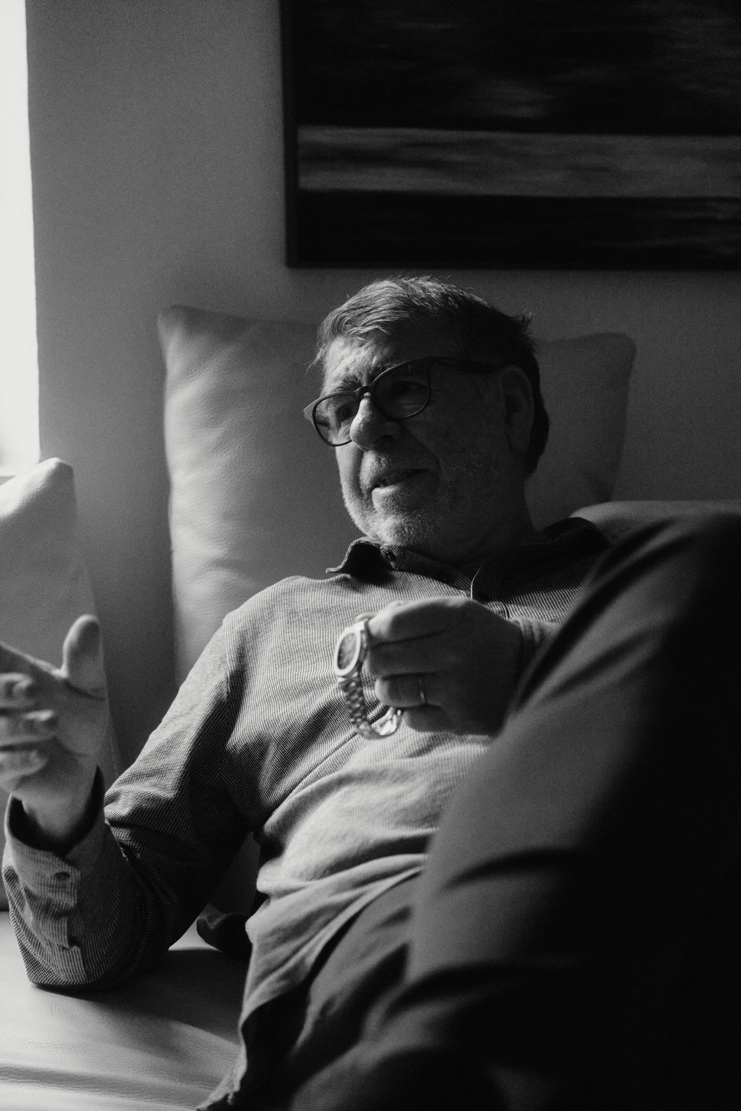 Ein Mann sitzt entspannt auf einem Sofa und hält eine Uhr in der Hand. Er trägt eine Brille und wirkt nachdenklich. Das Foto ist in Schwarz-Weiß gehalten.