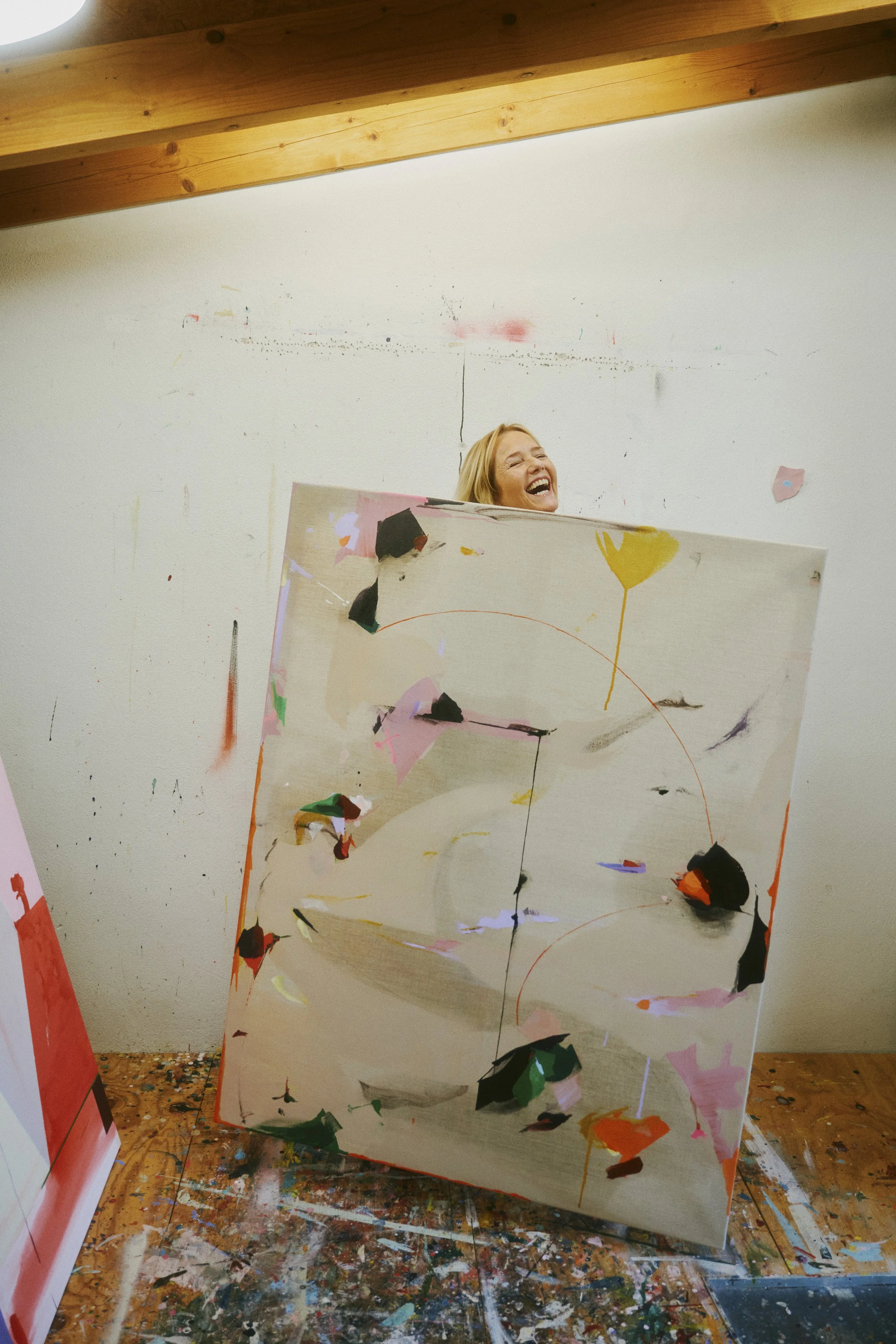 Lächelnde Person steht hinter einer abstrakten Leinwandmalerei in einem Atelier.