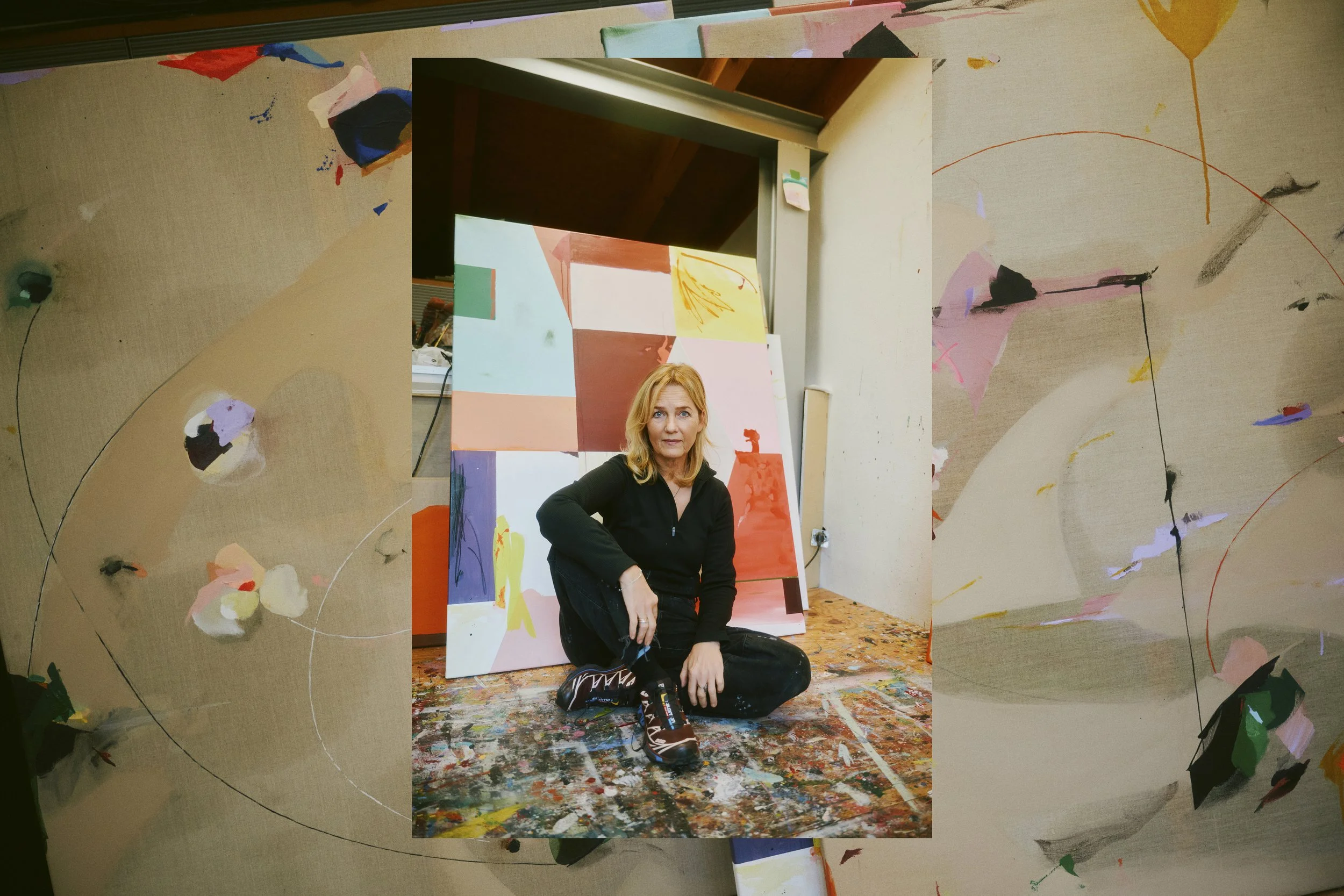 Eine Frau sitzt auf einem farbenfrohen Boden voller Farbflecken vor abstrakten Gemälden in einem Atelier. Sie trägt eine dunkle Kleidung und hat mittellanges blondes Haar. Die umgebenden Leinwände zeigen moderne, abstrakte Kunst mit verschiedenen Far