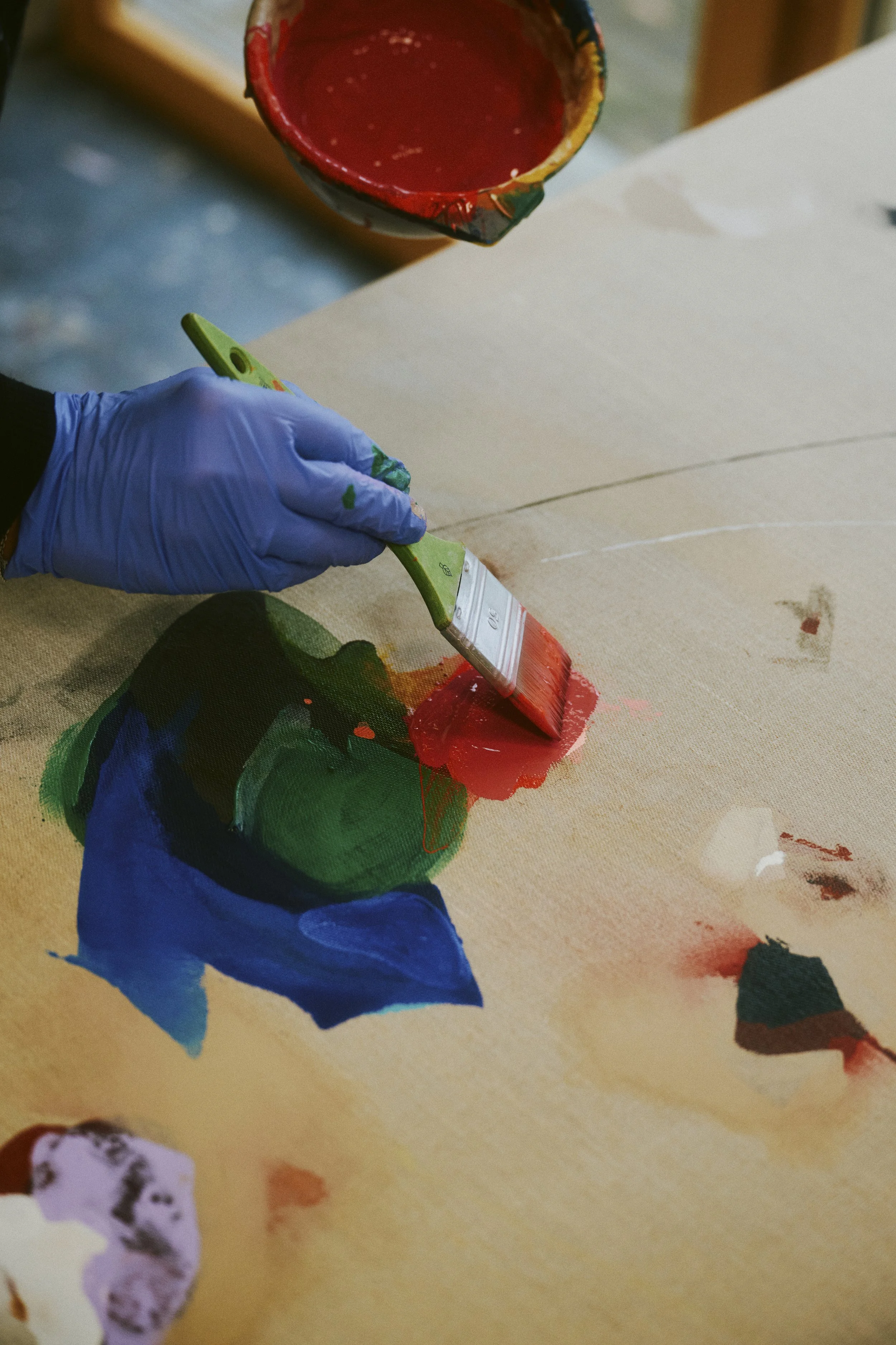Eine Person trägt mit einem Pinsel rote Farbe auf eine Leinwand auf. Die Hand trägt einen blauen Handschuh. Auf der Leinwand sind bereits andere Farben wie Blau und Grün sichtbar.