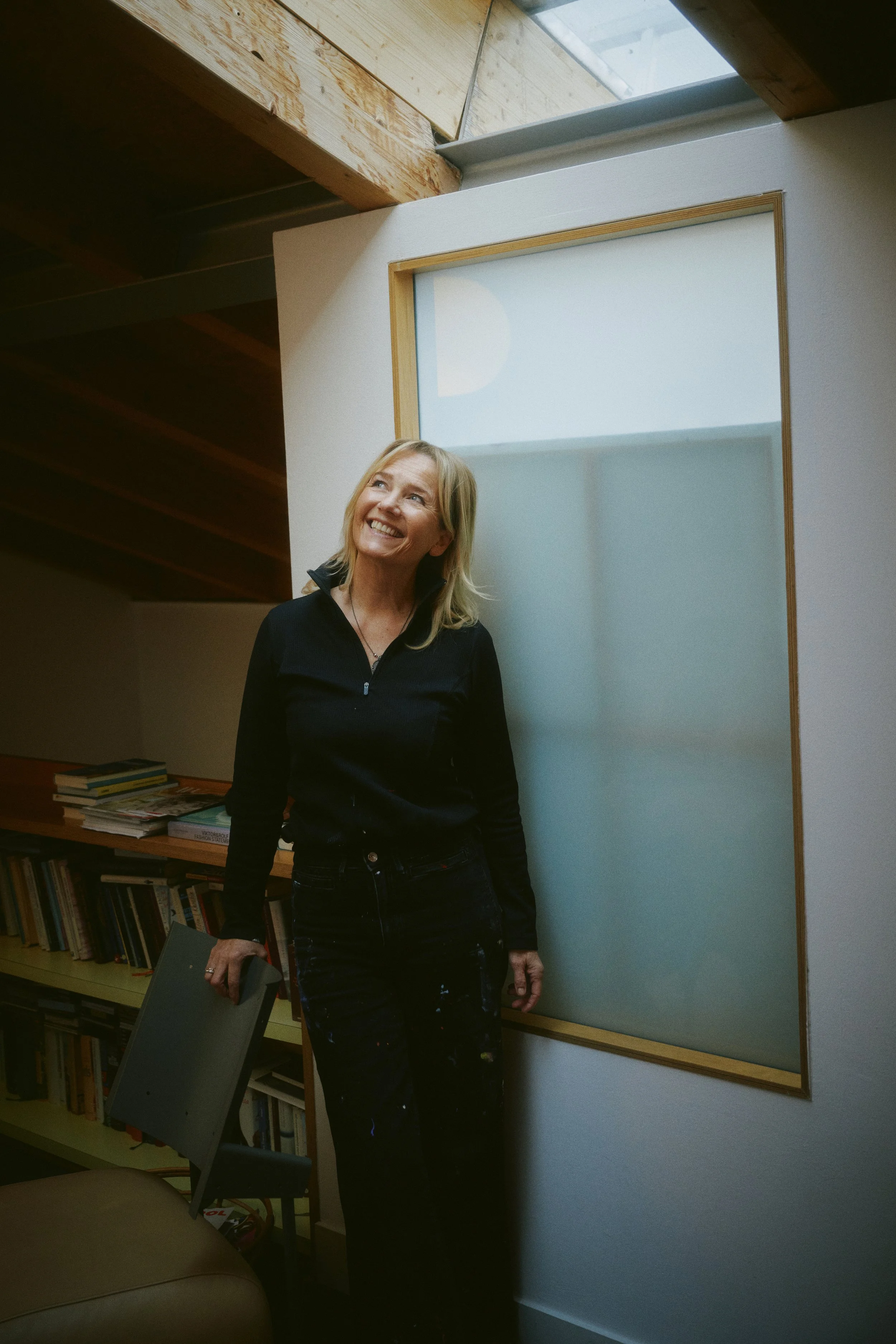 Eine lächelnde Frau in einem dunklen Raum mit Bücherregalen im Hintergrund, die neben einem Glasfenster steht.