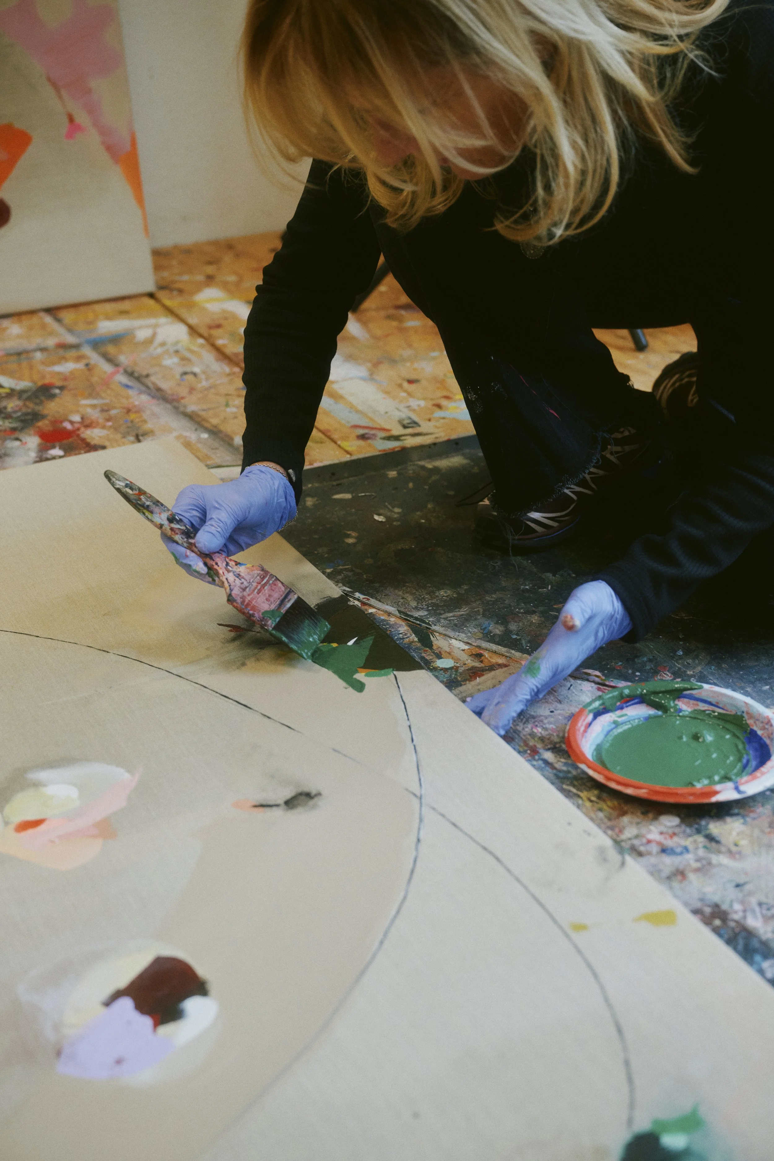 Künstler arbeitet auf Leinwand mit grüner Farbe; trägt Handschuhe; umgeben von buntem Atelierboden.