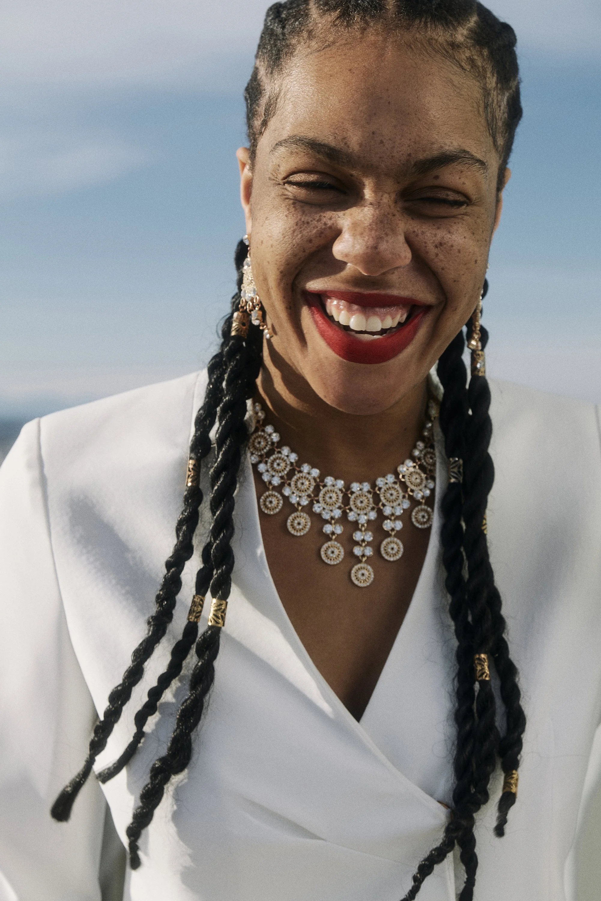 Eine lachende QUEEN Lizzy mit langen Zöpfen und auffälligem Schmuck in einem weißen Kleid vor blauem Himmel.