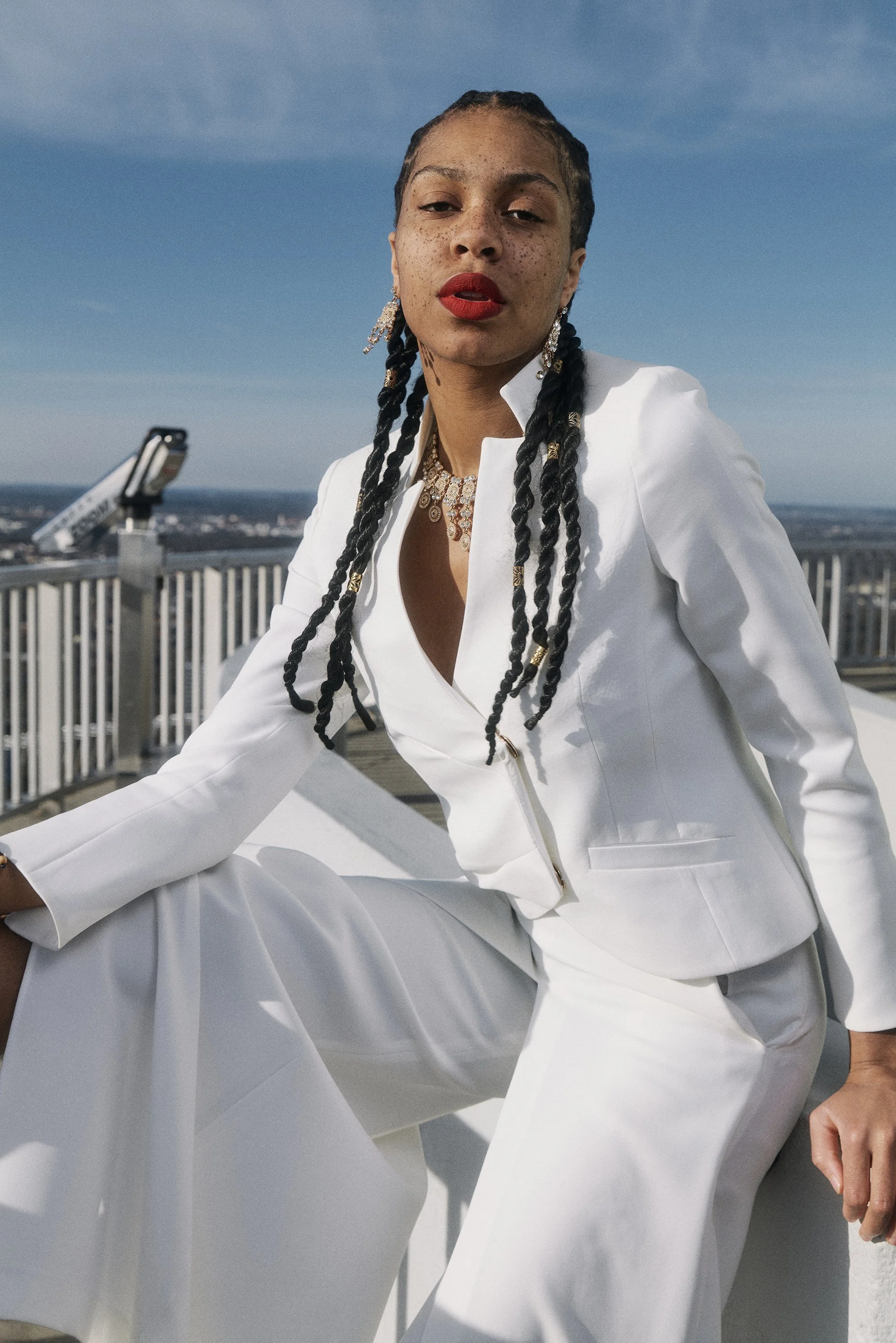 QUEEN Lizzy in weißem Anzug mit langen Zöpfen, rotem Lippenstift und Schmuck, sitzt auf einem Dach vor einem Geländer, Himmel im Hintergrund.