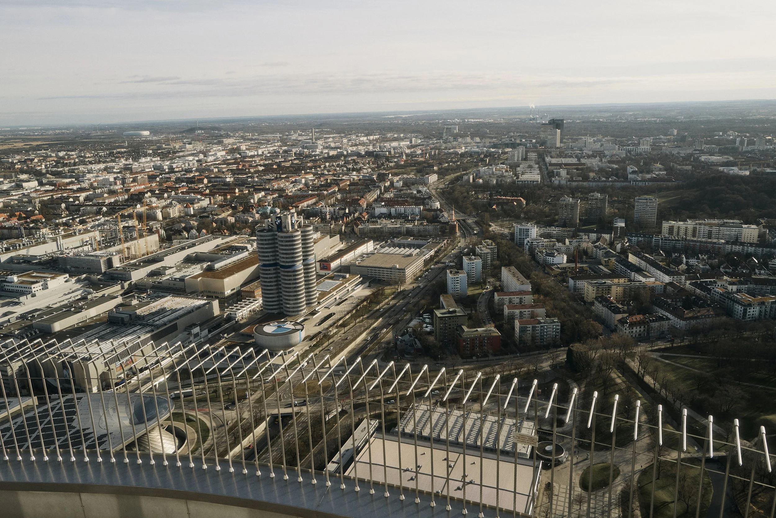 Blick auf eine Stadt mit modernen Gebäuden und einem markanten runden Turm, umgeben von einem weiten urbanen Gebiet, aufgenommen aus der Vogelperspektive.
