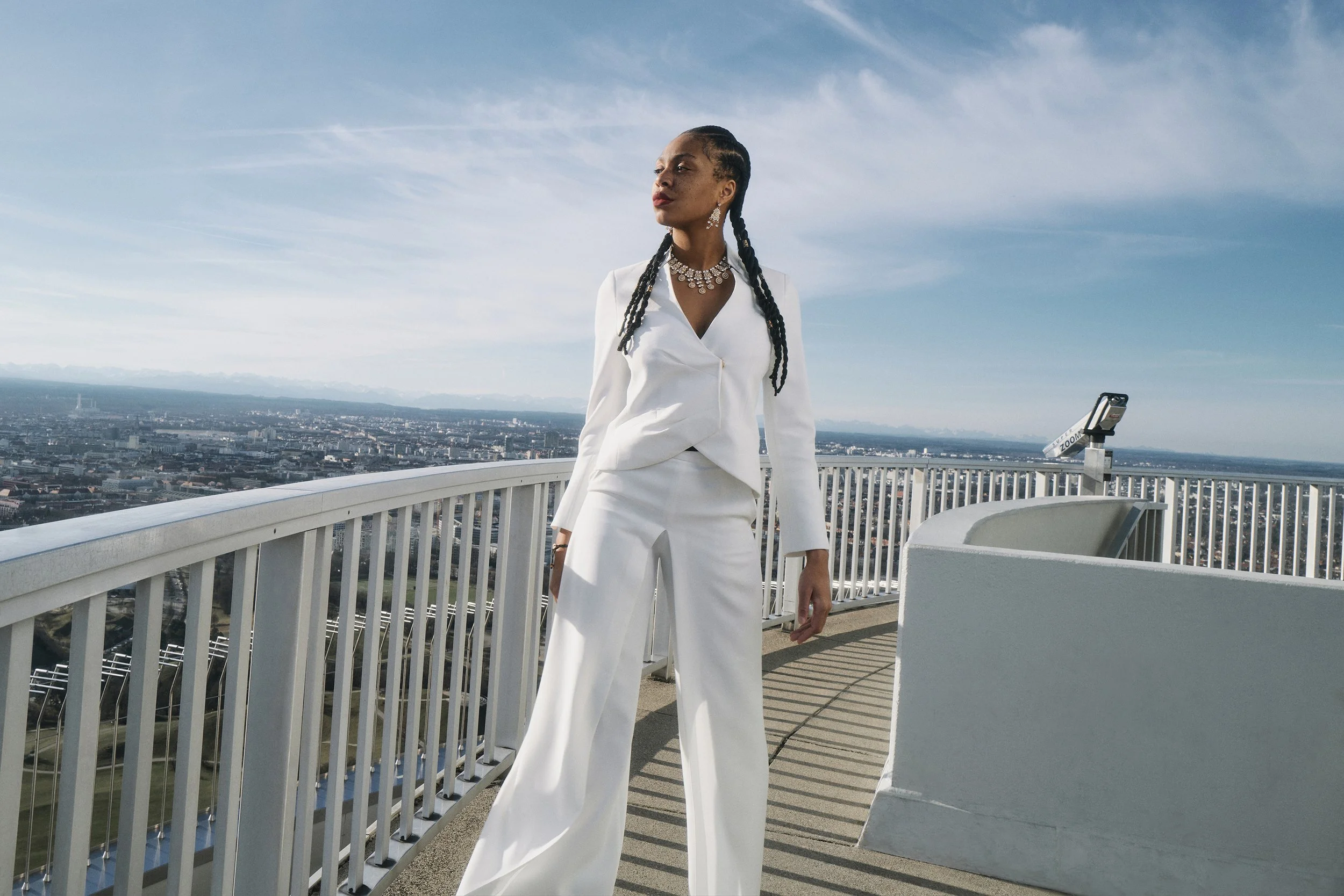 QUEEN Lizzy in einem weißen Anzug steht auf einer Aussichtsplattform. Im Hintergrund ist eine Stadtlandschaft unter einem blauen Himmel zu sehen. Die Frau hat lange geflochtene Haare und trägt Schmuck.