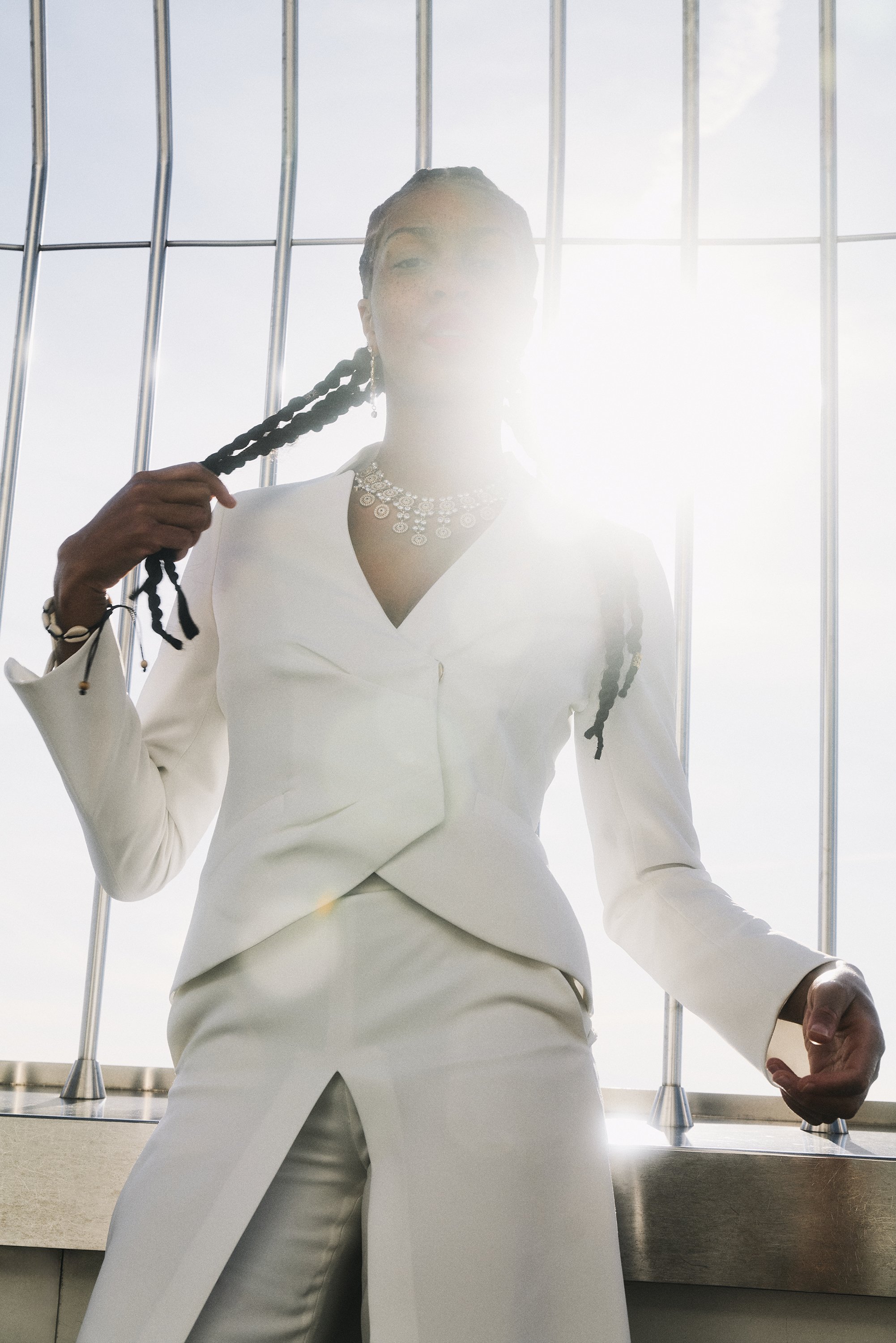 Person in weißem Anzug mit geflochtenem Haar und Schmuck vor einem hellen Hintergrund fotografiert.