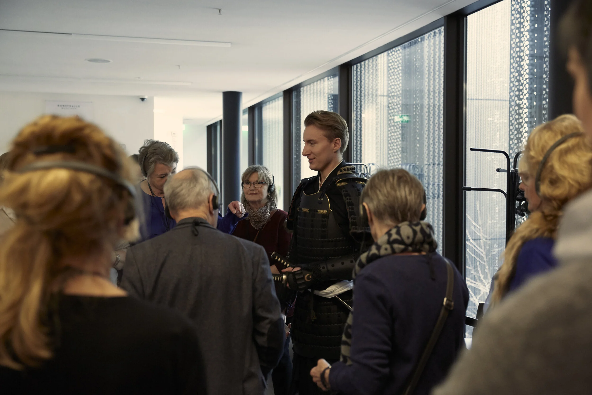 Menschengruppe in einem modernen Raum, eine Person in traditioneller Samurai-Rüstung umgeben von anderen Personen, die Kopfhörer tragen.