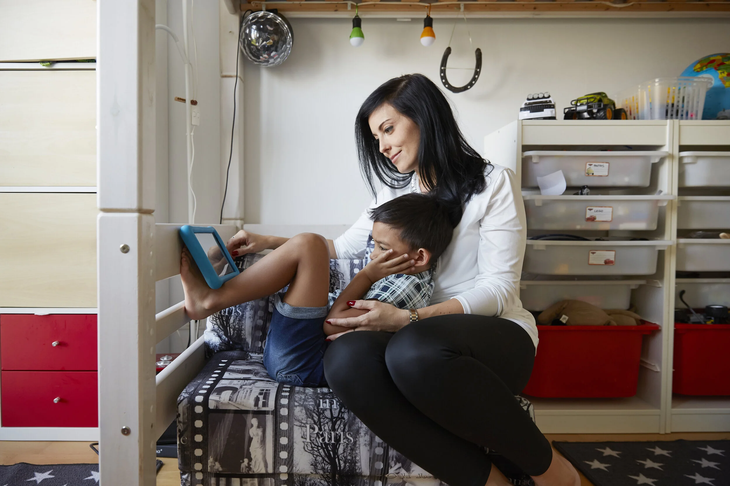 Eine Frau und ein Kind sitzen in einem Kinderzimmer. Das Kind sitzt auf dem Schoß der Frau und hält ein Tablet mit den Füßen. Im Hintergrund sind Regale mit Spielzeug und ein Etagenbett zu sehen.
