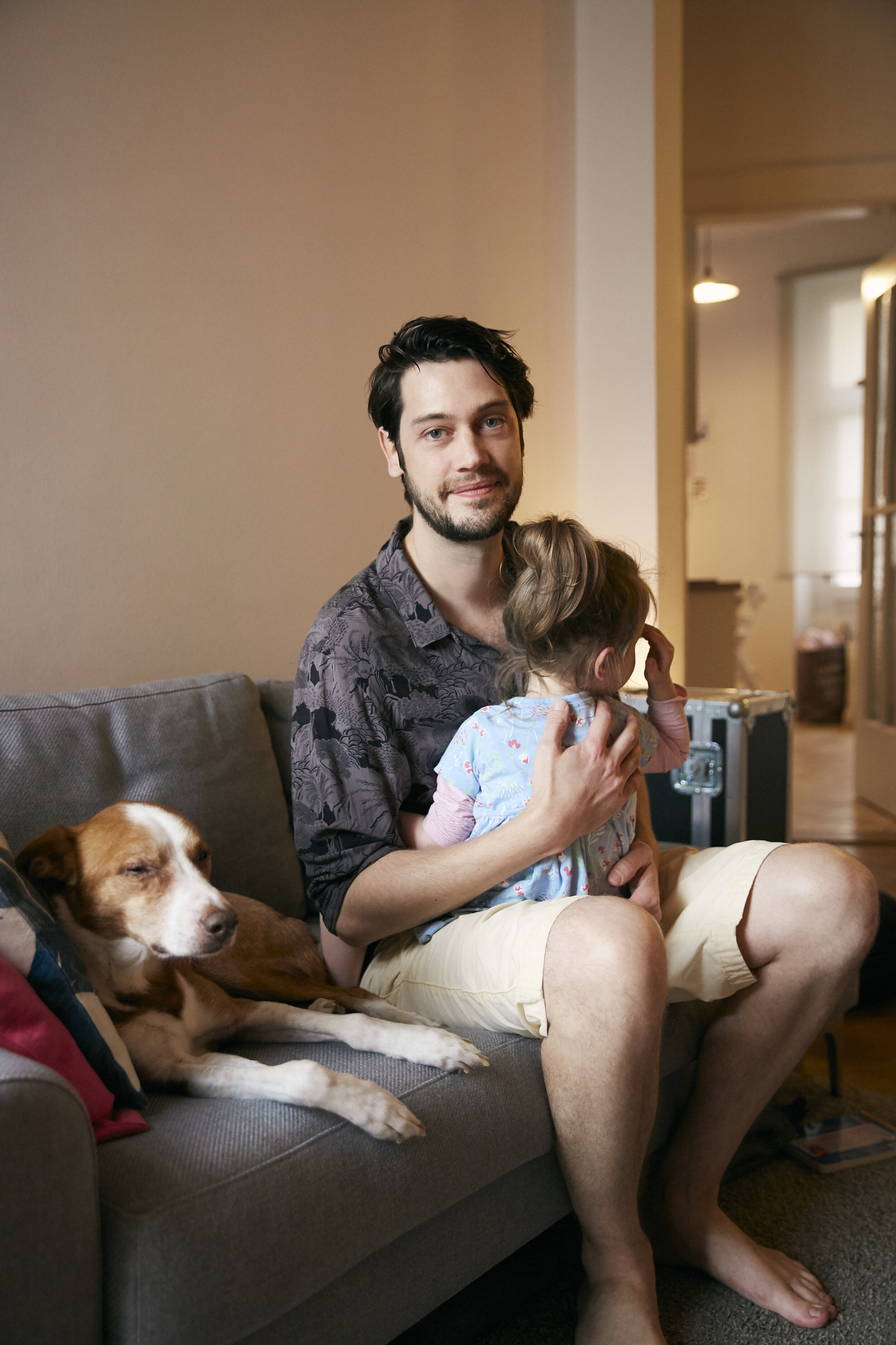 Ein Mann sitzt auf einem Sofa, hält ein Kind im Arm und ein Hund liegt neben ihnen. Der Raum hat eine warme Beleuchtung.
