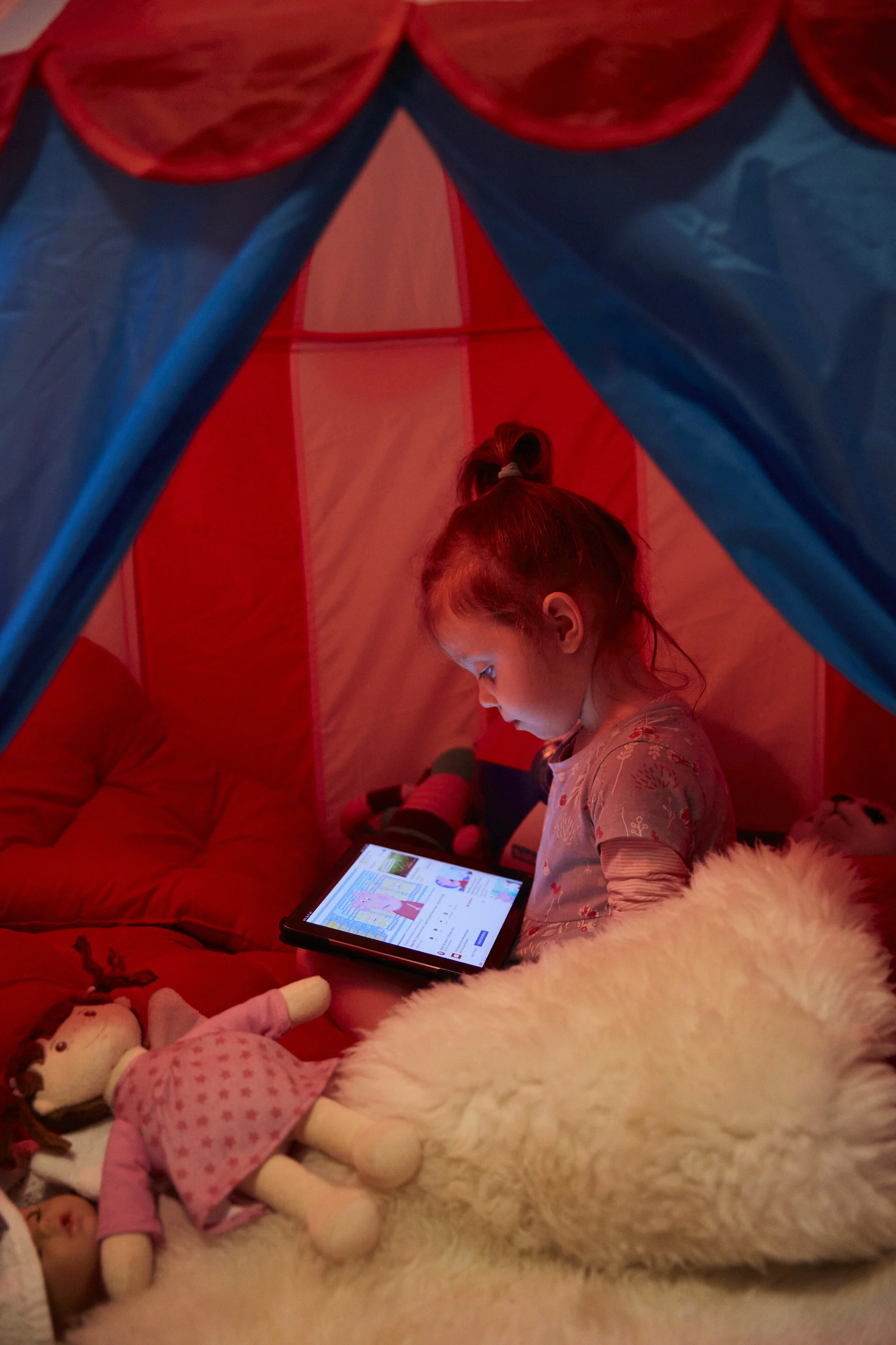 Mädchen sitzt in einem Zelt und schaut auf ein Tablet, umgeben von Kuscheltieren und Puppen.