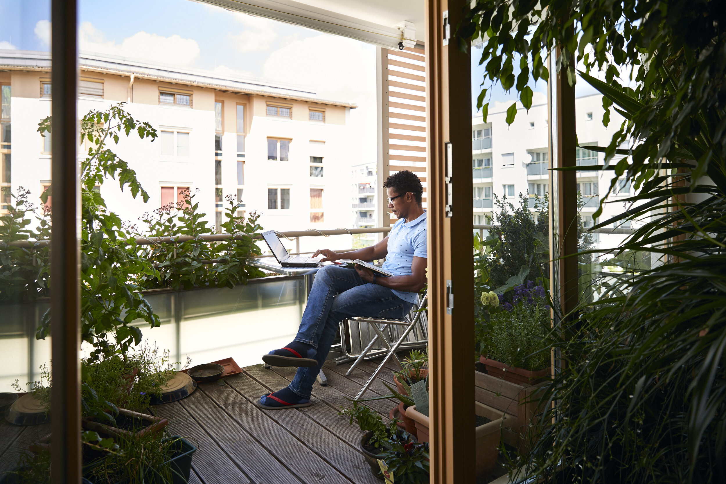 Mann sitzt auf einem Balkon, arbeitet mit Laptop, umgeben von Pflanzen, moderner städtischer Hintergrund.