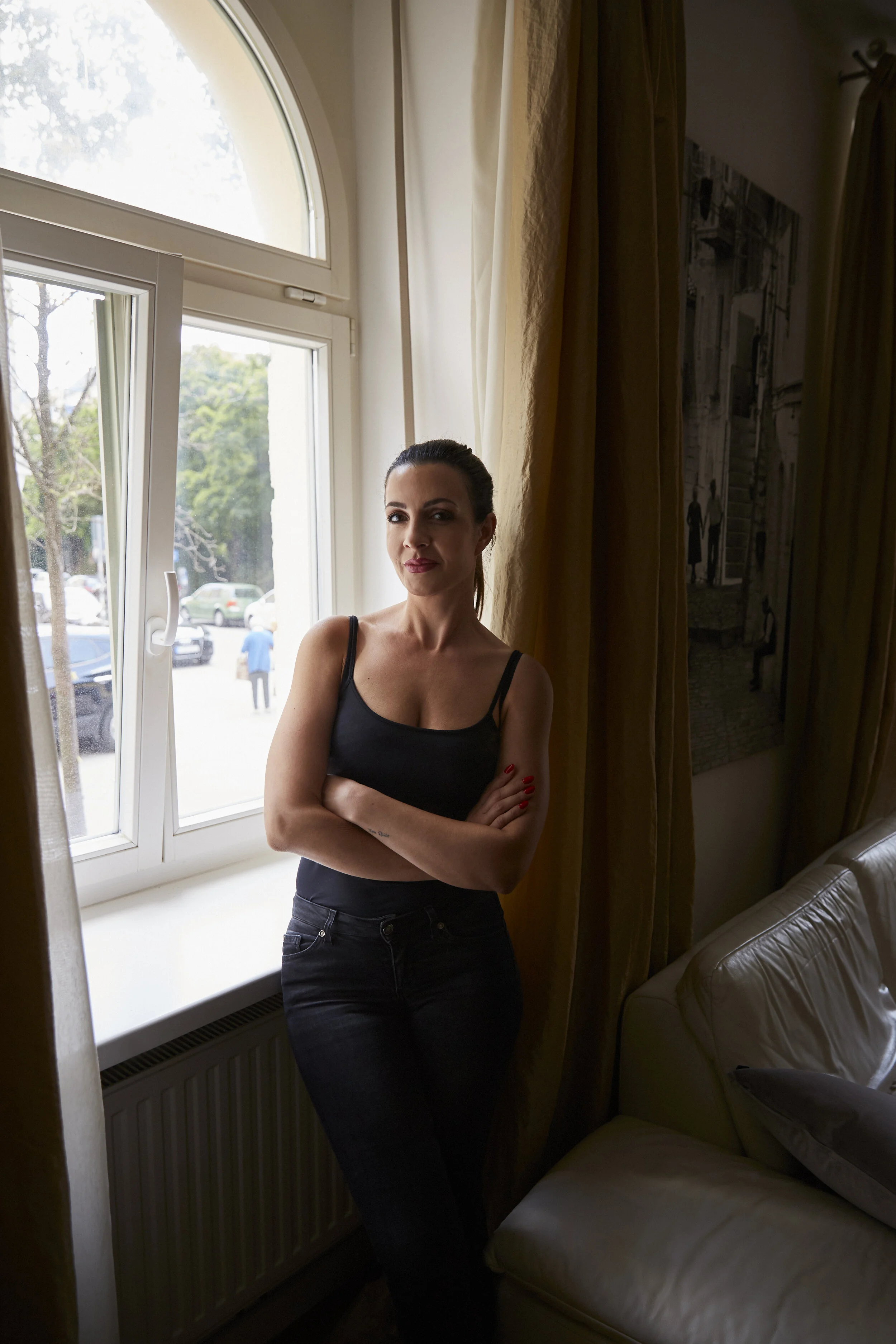 Frau in schwarzem Top steht mit verschränkten Armen am Fenster in einem Zimmer; draußen sind Autos und Menschen zu sehen.