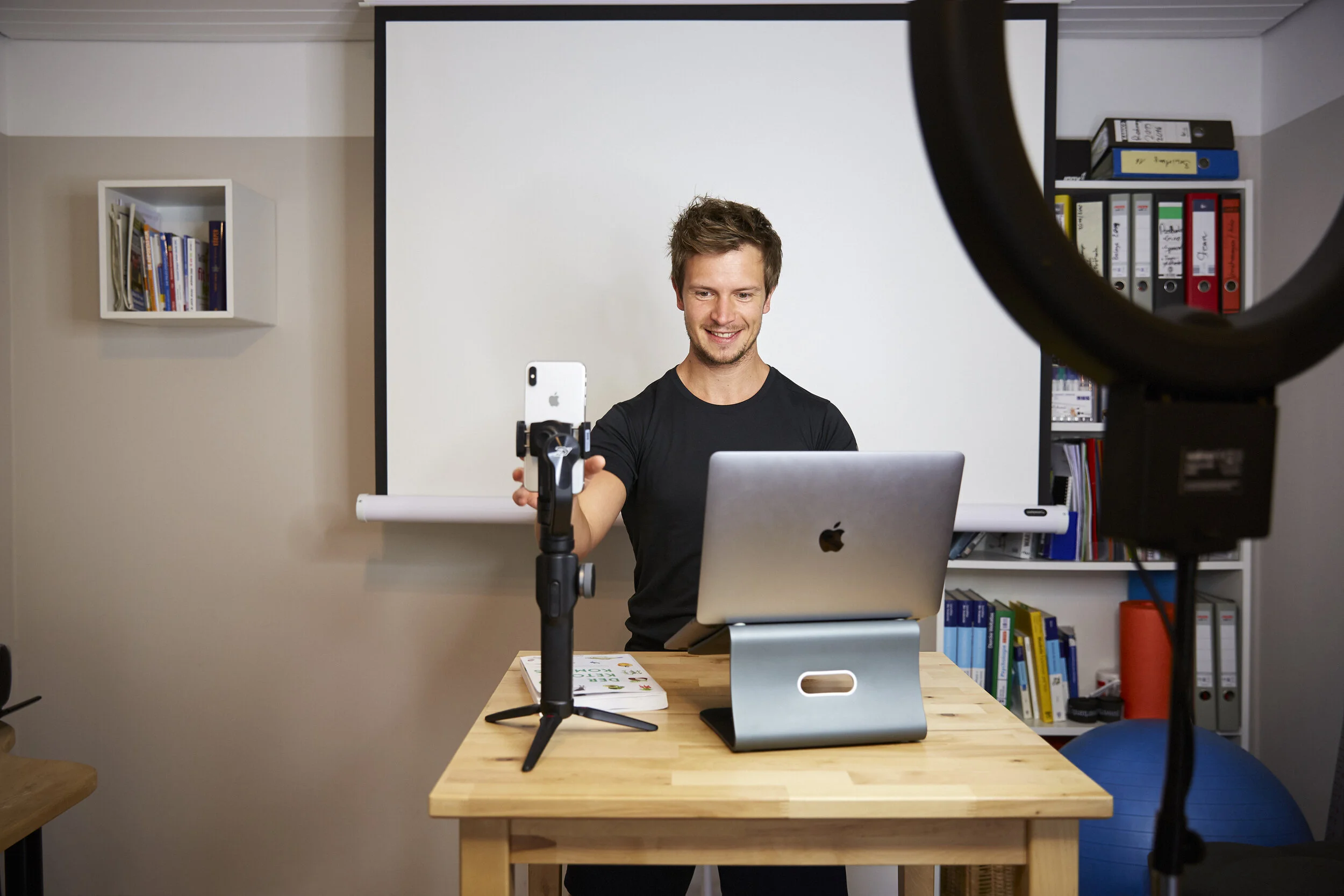 Ein Mann sitzt an einem Tisch mit einem Laptop und einem Smartphone auf einem Stativ. Im Hintergrund hängt ein Bücherregal. Ein Ringlicht ist eingestellt, um die Szene zu beleuchten.