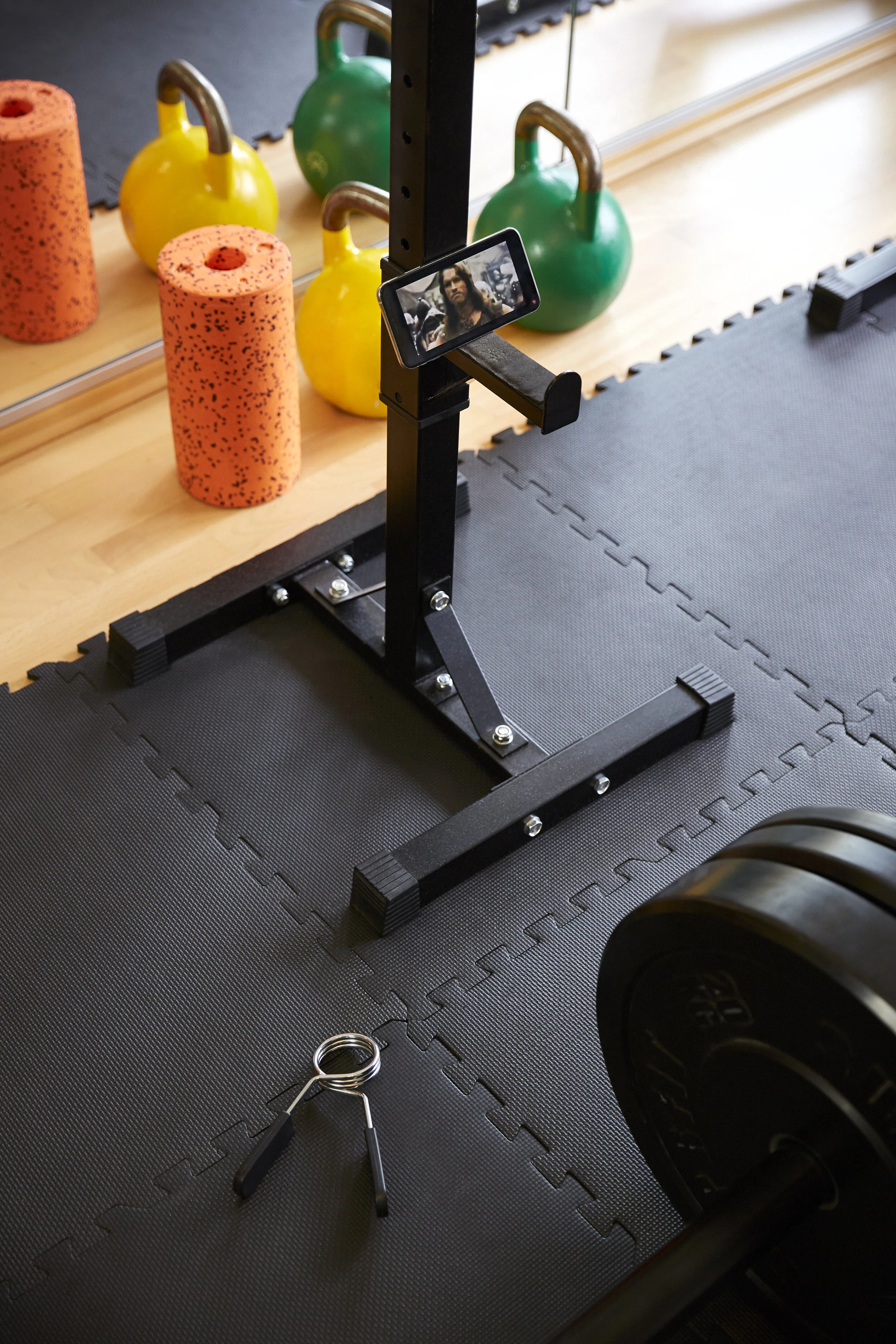 Heim-Fitnessstudio mit Kettlebells, Schaumstoffrollen, Smartphone, Hantel, Gewichtsscheiben und Federklemme auf Gummimatte.