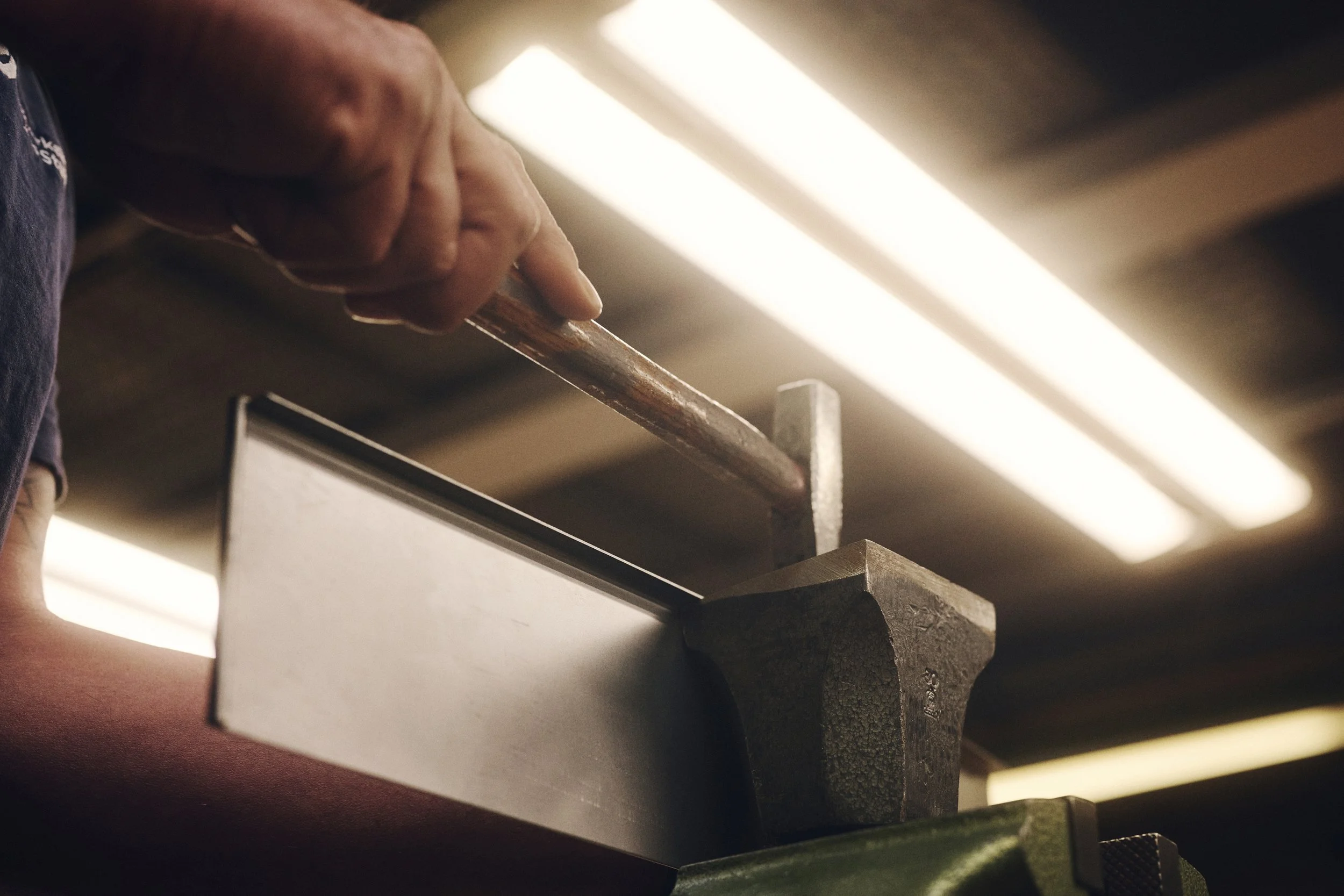 Nahaufnahme einer Hand, die in einer Werkstatt mit einem Hammer auf ein Metallstück schlägt.