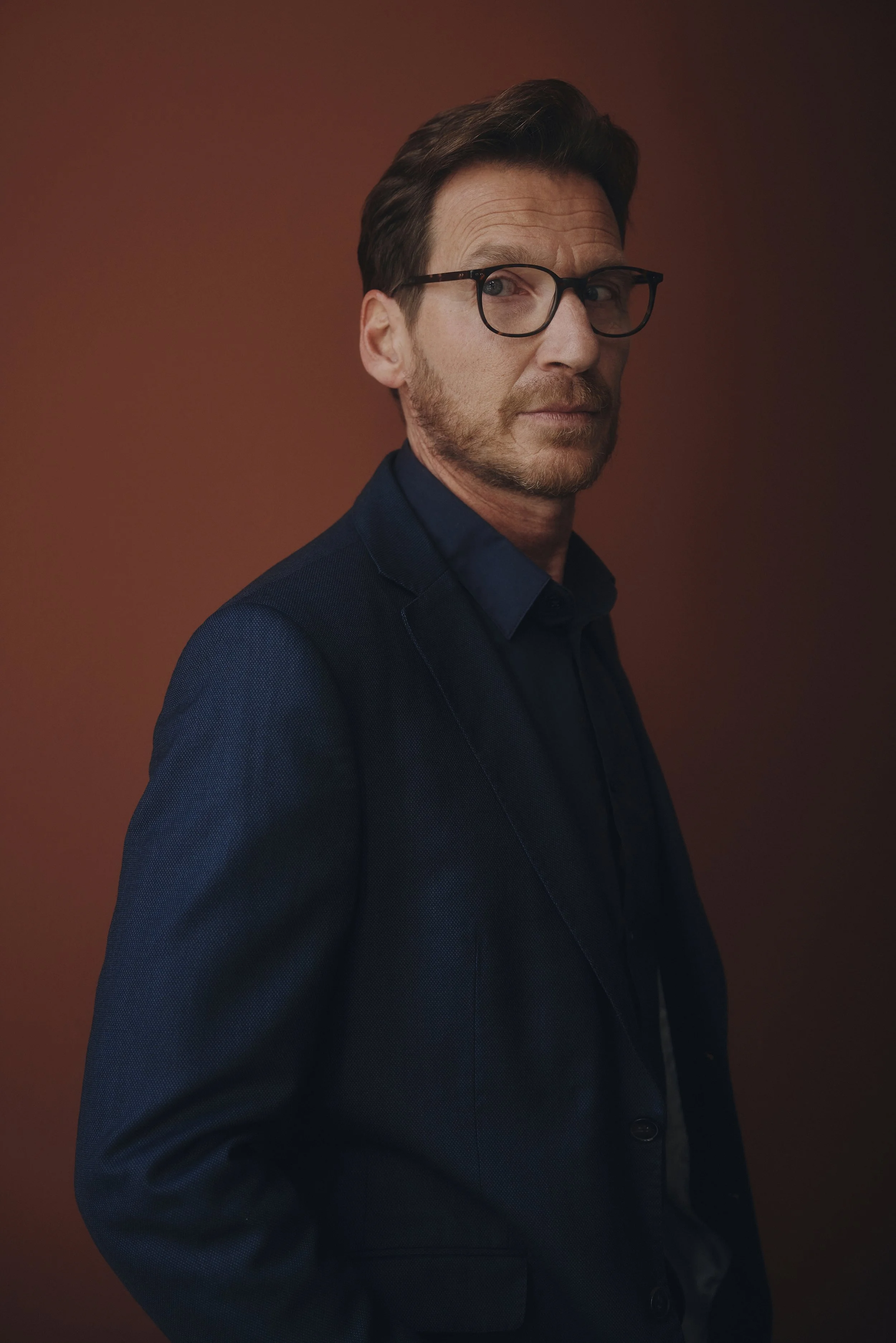 Mann mit Brille und Anzug vor einem braunen Hintergrund.