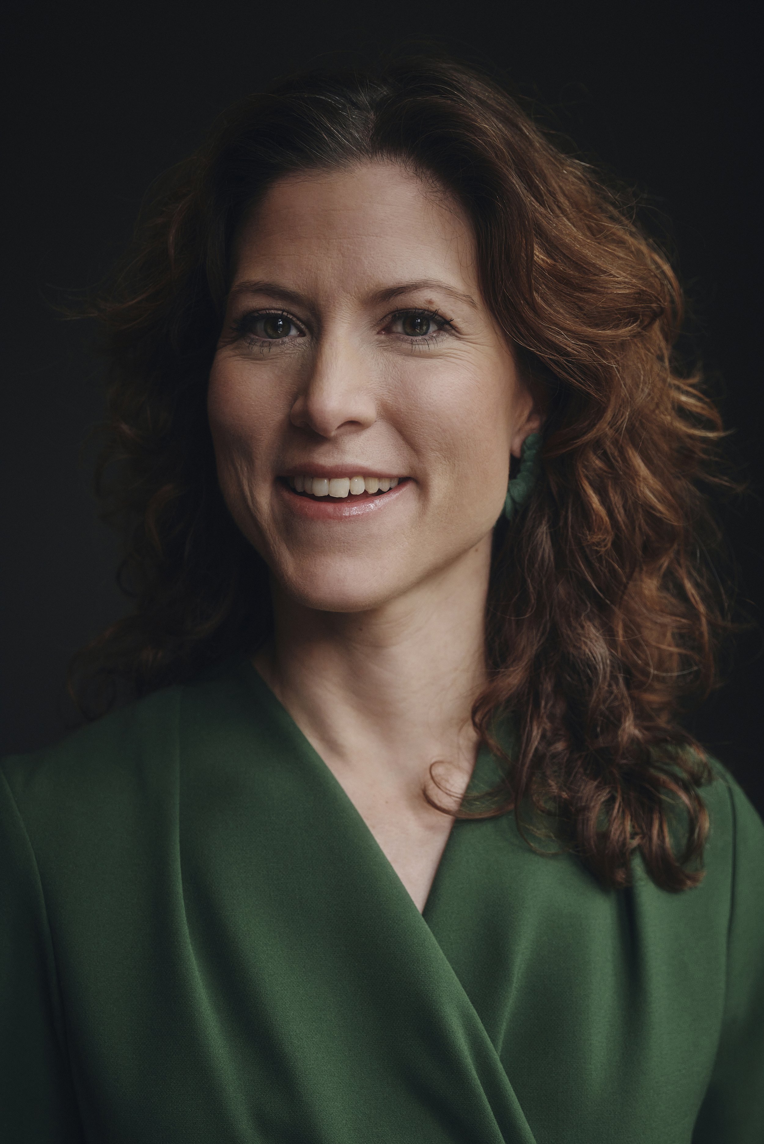 Porträt einer Frau mit lockigem Haar, die ein grünes Oberteil trägt und lächelt, vor einem dunklen Hintergrund.