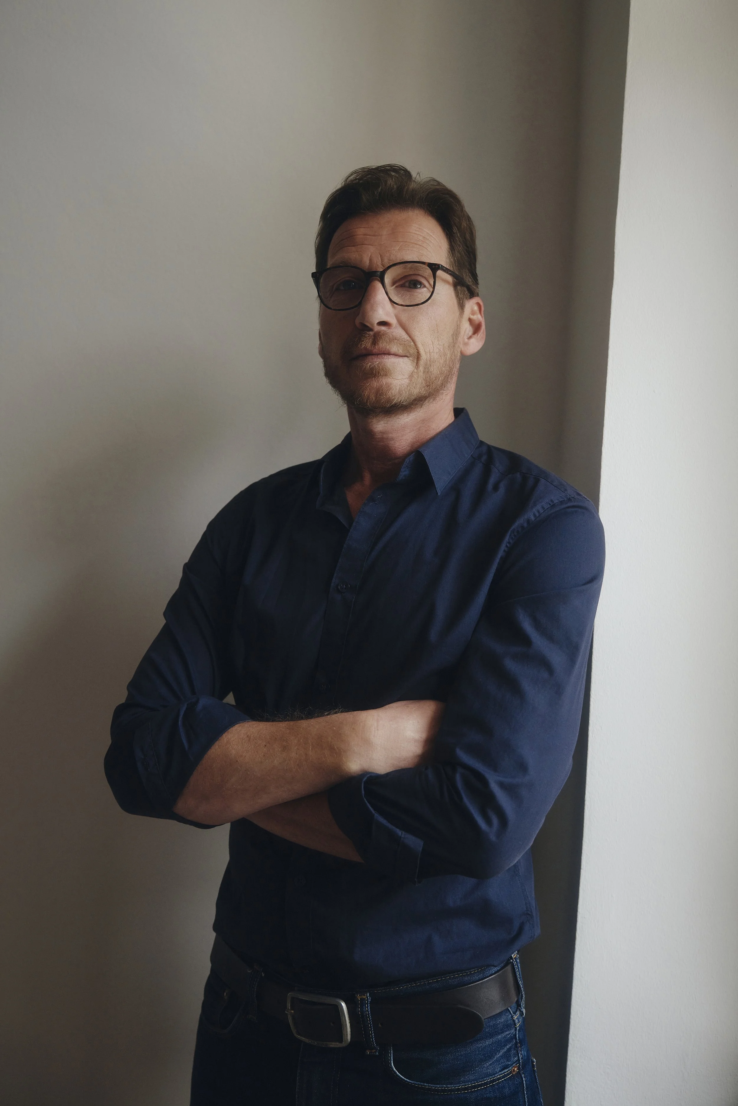 Mann mit Brille, blauem Hemd, verschränkten Armen, steht vor einer weißen Wand.