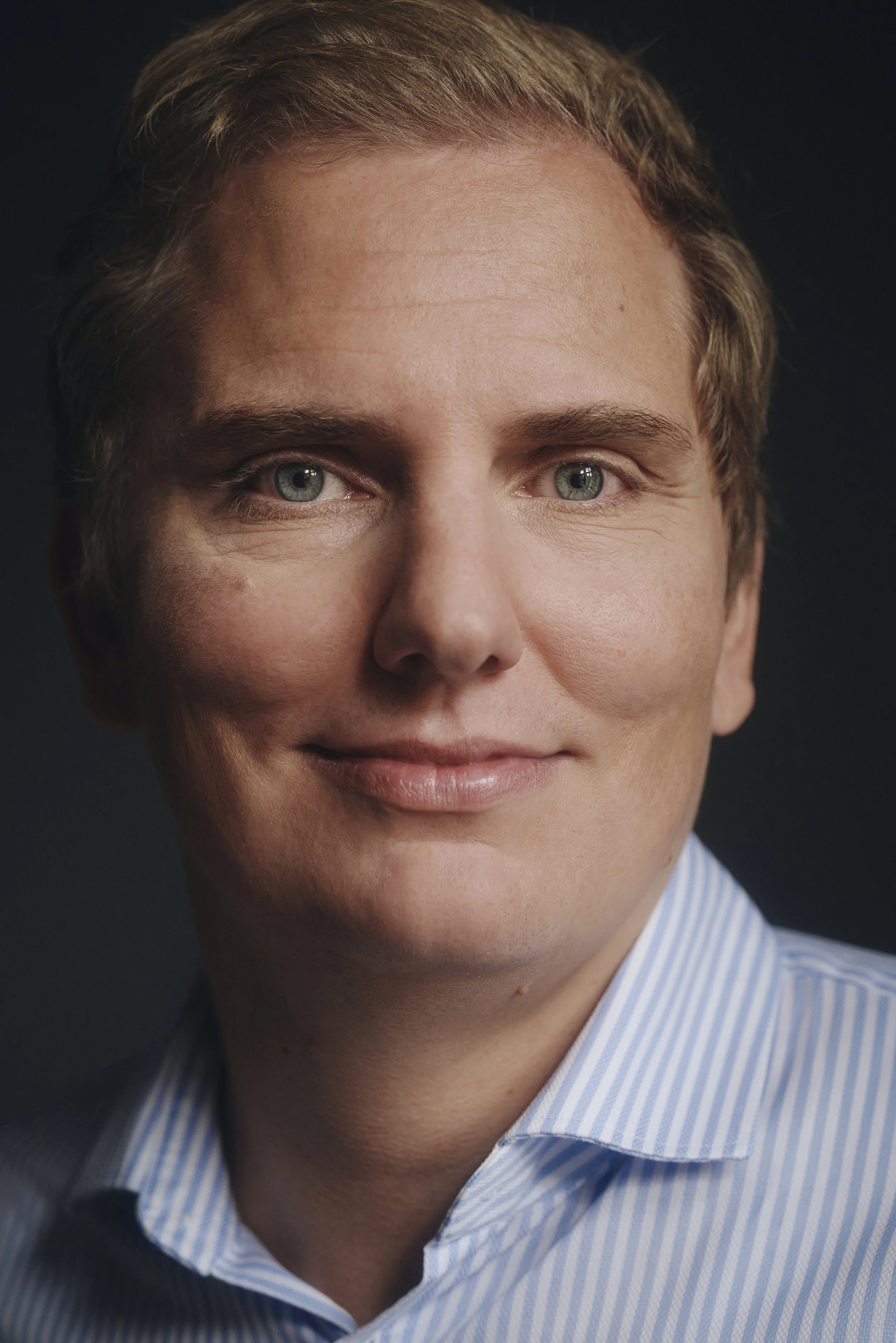 Porträt eines Mannes mit blauer gestreifter Hemd, lächelnd, vor dunklem Hintergrund.