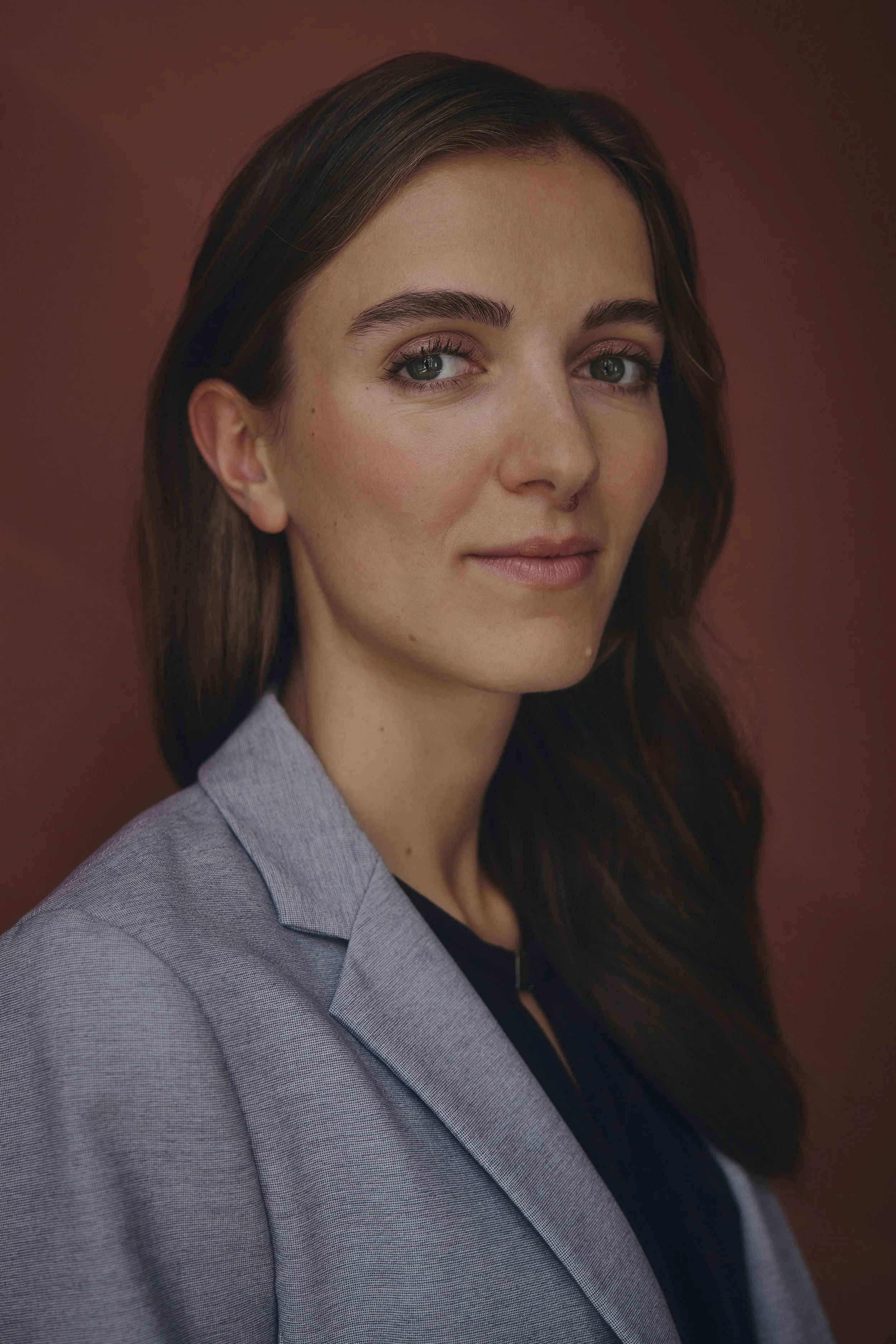 Porträt einer Frau mit braunen Haaren, die einen grauen Blazer trägt, vor einem rotbraunen Hintergrund.