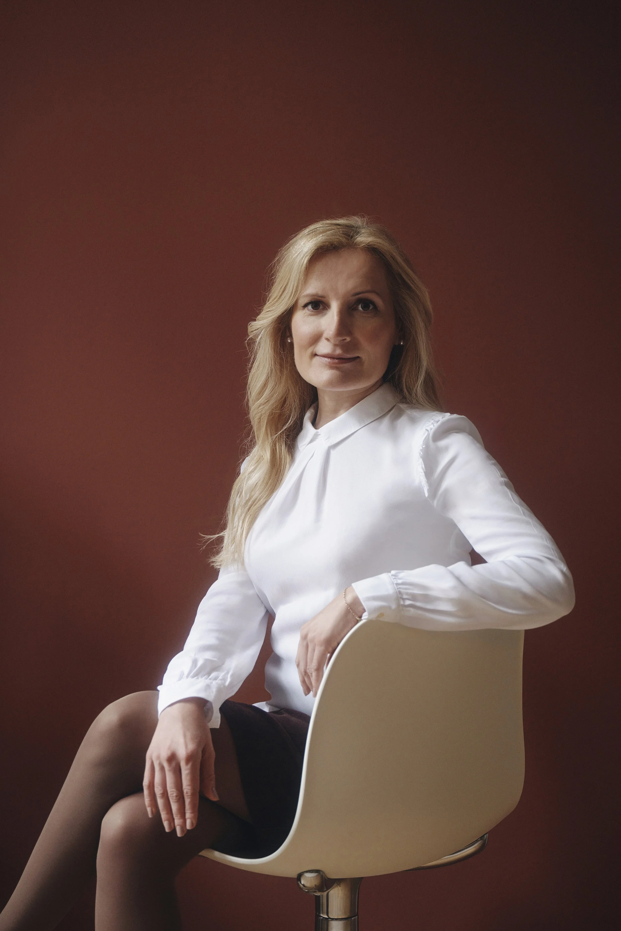 Frau sitzt auf einem Stuhl vor einem dunklen Hintergrund, trägt eine weiße Bluse und blickt in die Kamera.