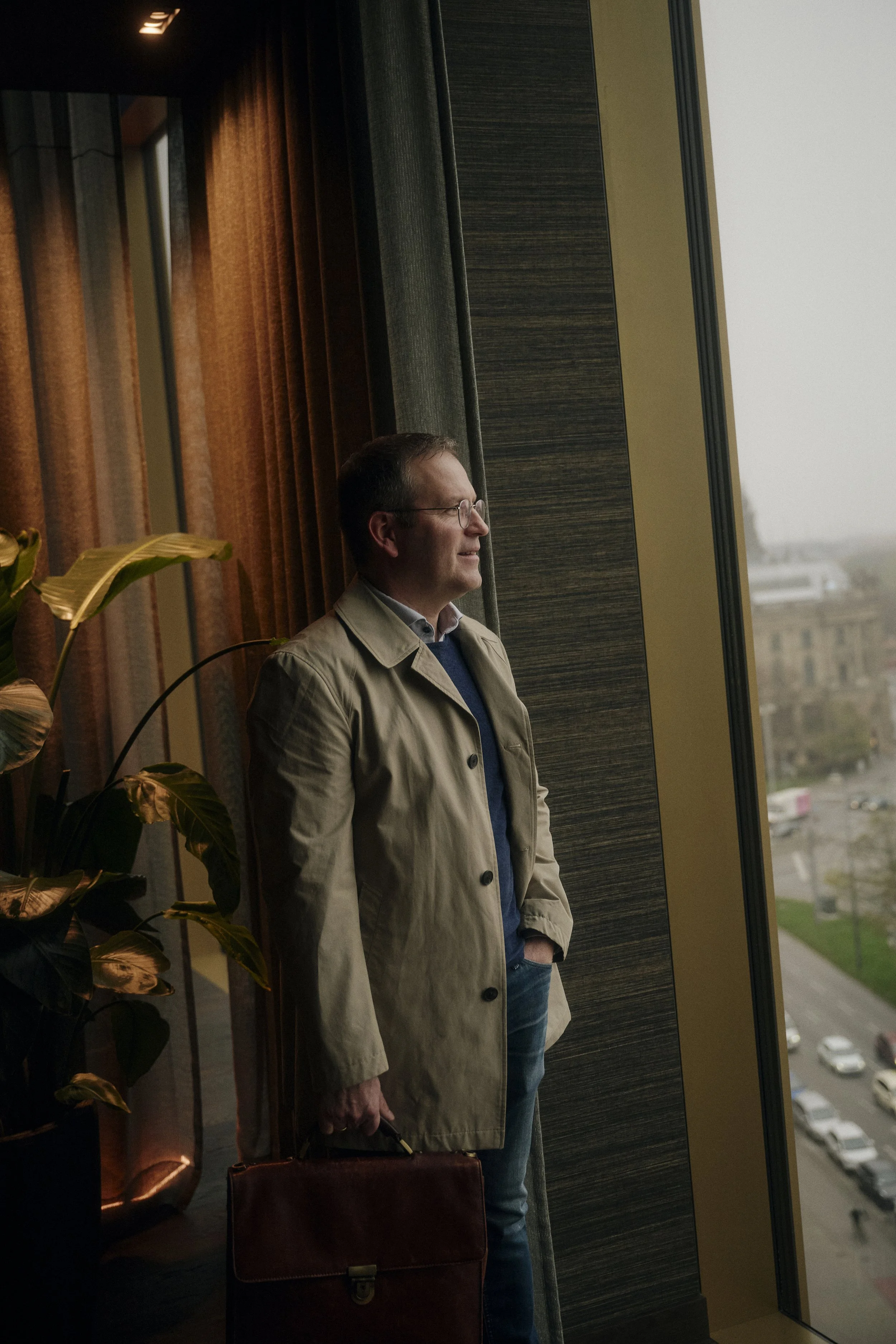 Mann mit Brille in einem Beigefarbenen Mantel steht am Fenster und schaut hinaus. Er hält eine braune Aktentasche und steht in der Nähe von Pflanzen und grauen Vorhängen. Draußen sind verschwommene Gebäude und Straßen zu sehen.