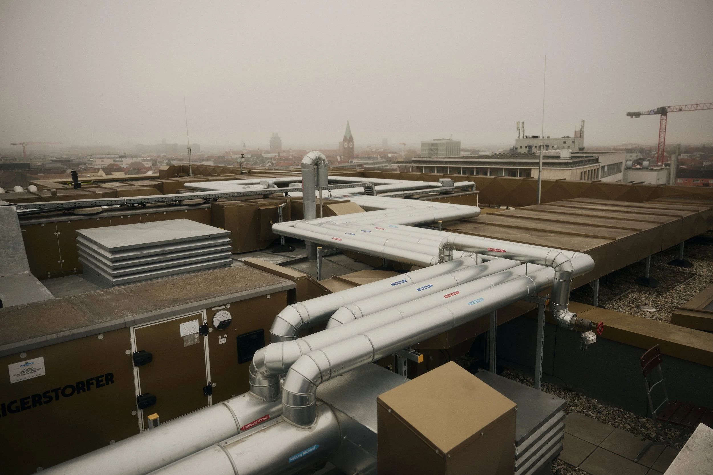Blick über eine städtische Dachlandschaft mit Lüftungsanlagen und Rohren, trübem Himmel im Hintergrund.